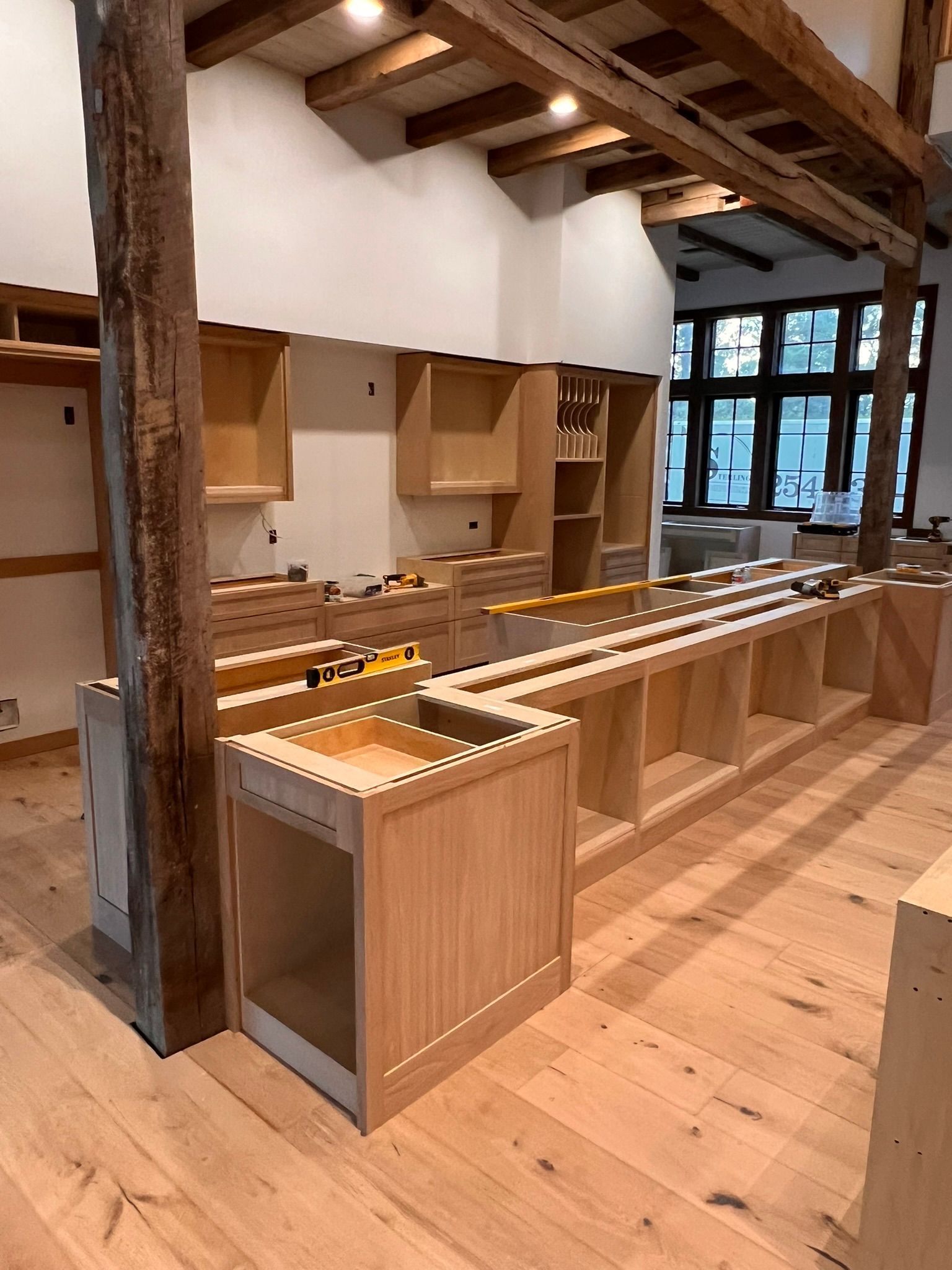 Kitchen under construction with unfinished light wood cabinets, wood beams, and hardwood floors.