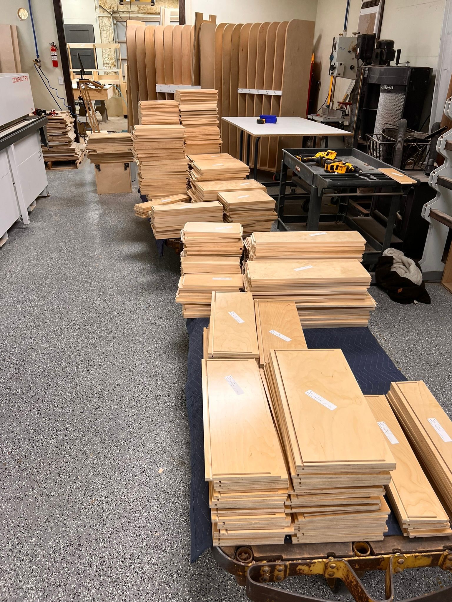 Stacks of light wood cabinet parts on a shop floor.  Various other wood pieces and work tables are in the background.