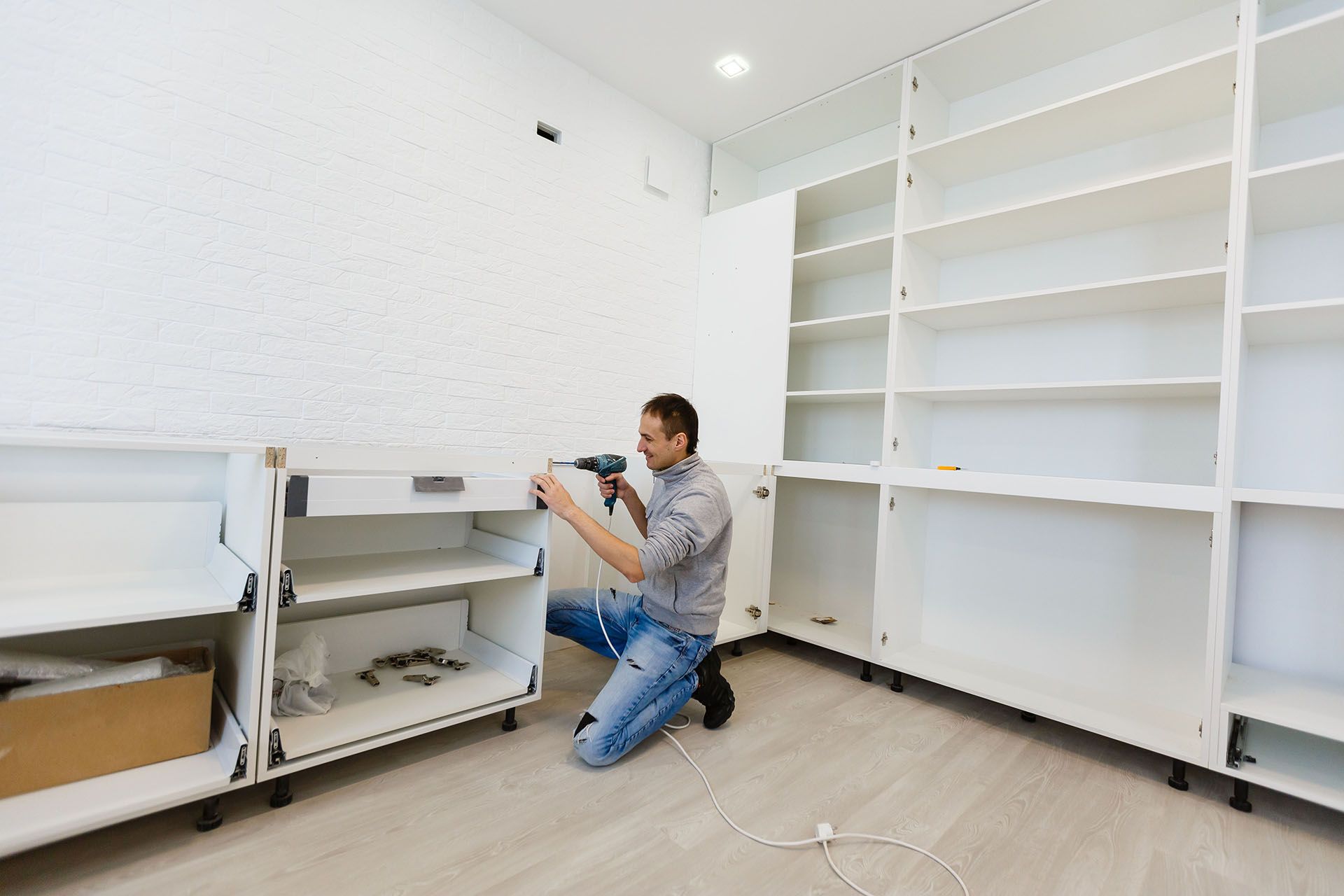 Person kneeling in a bright room, installing white commercial shelving units near the wall.