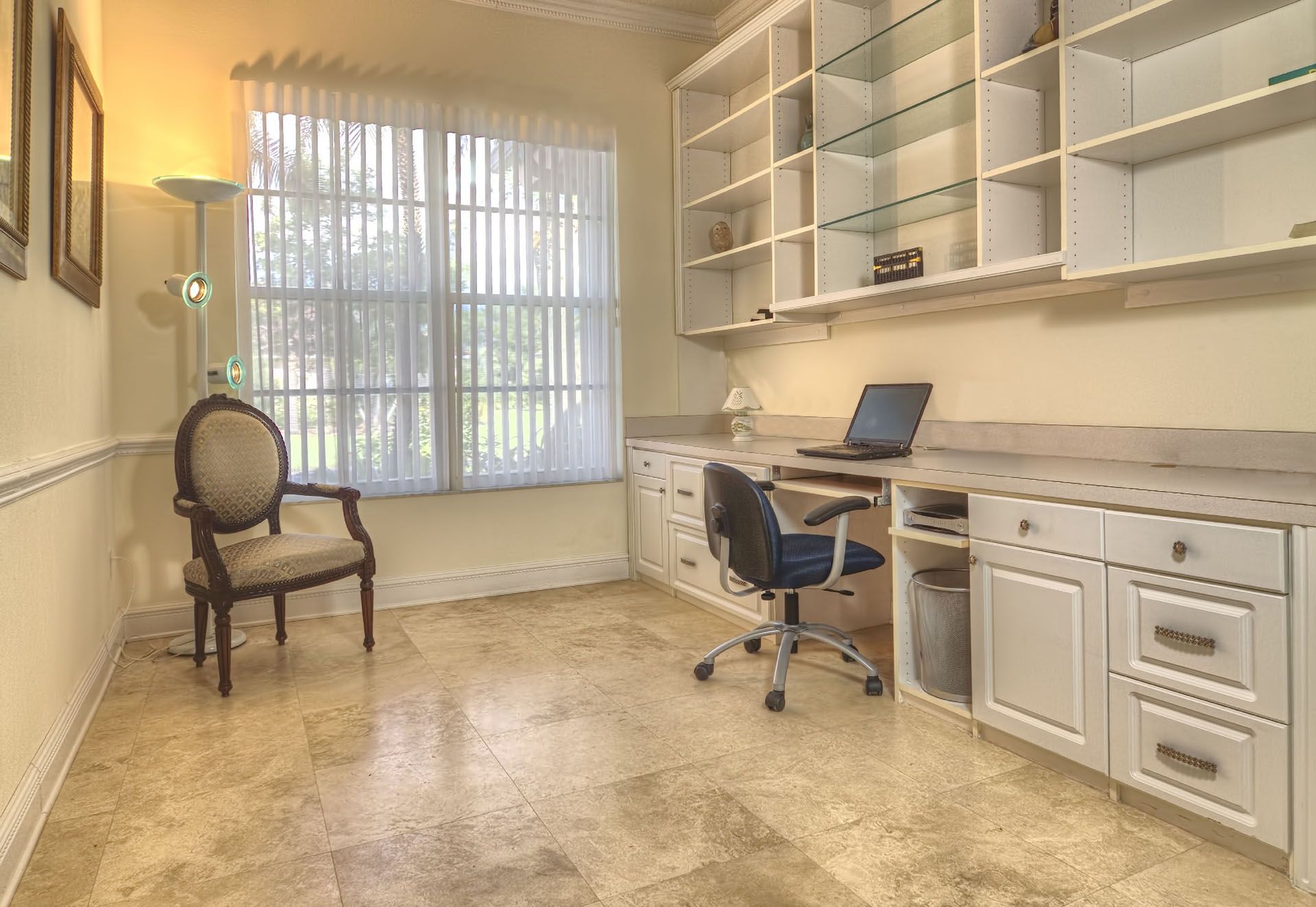 Bright home office with desk, laptop, shelves, chair, and a large window with blinds