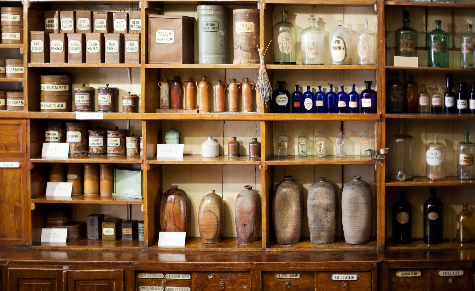 Wooden pharmacy shelves filled with vintage bottles, jars, and boxes in a warm, old-fashioned shop.