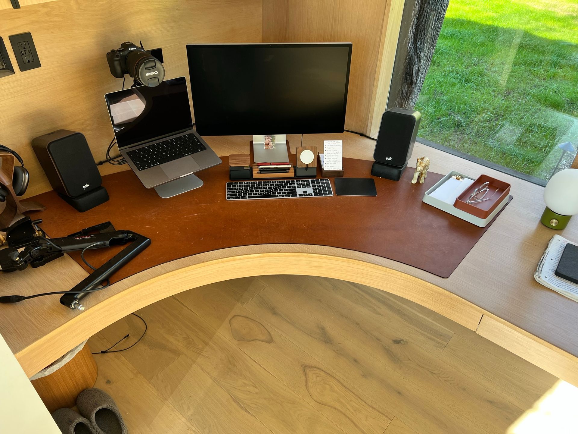 Wooden desk with multiple screens and tech equipment in front of a window, with a view of a green lawn.