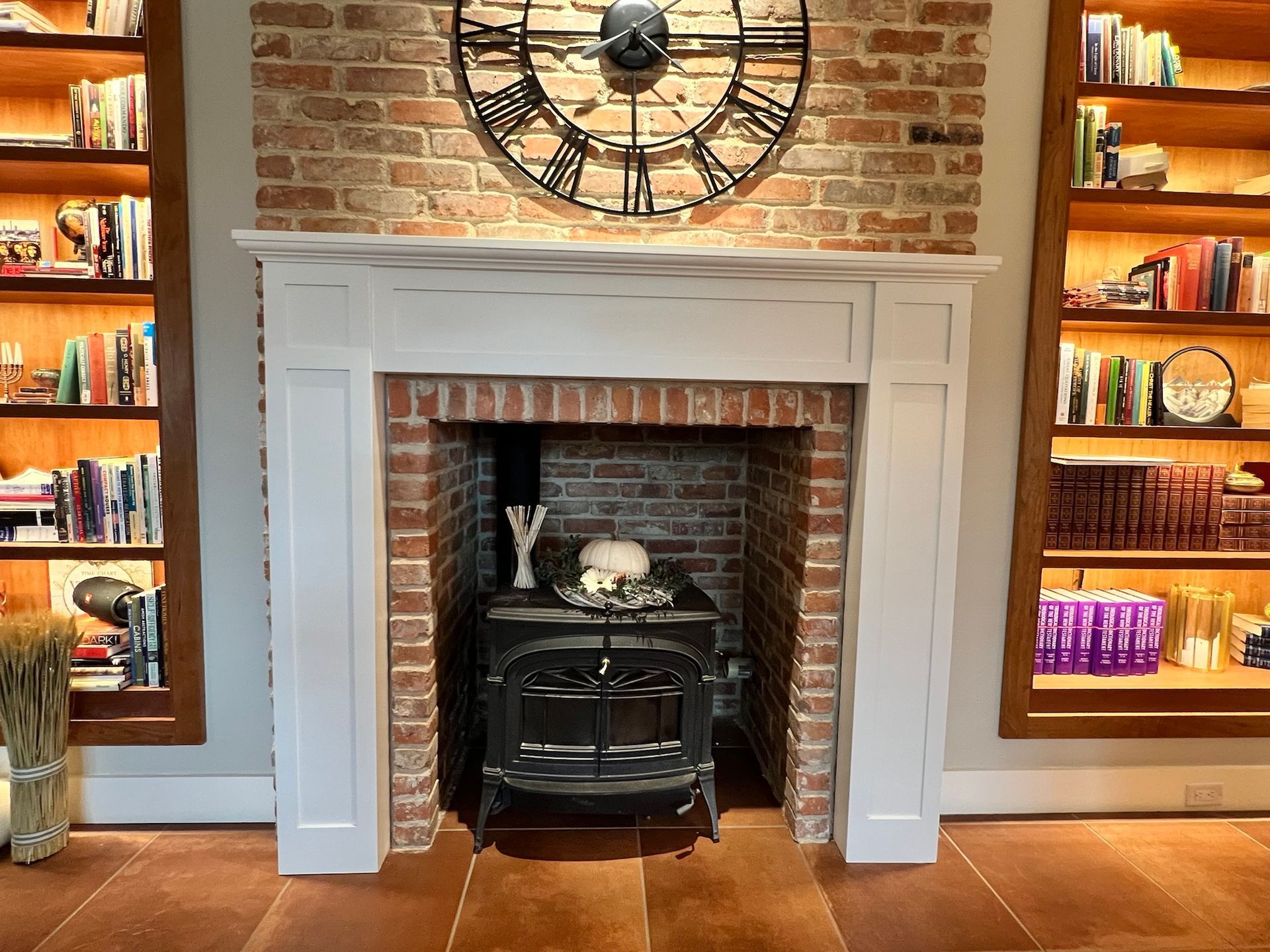Fireplace with brick interior, white mantle, and large clock. Bookshelves flank sides.