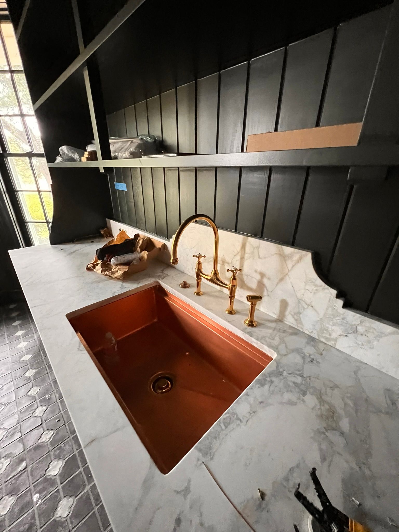 Copper sink in a marble countertop, gold faucet, black shelving, and dark paneling.