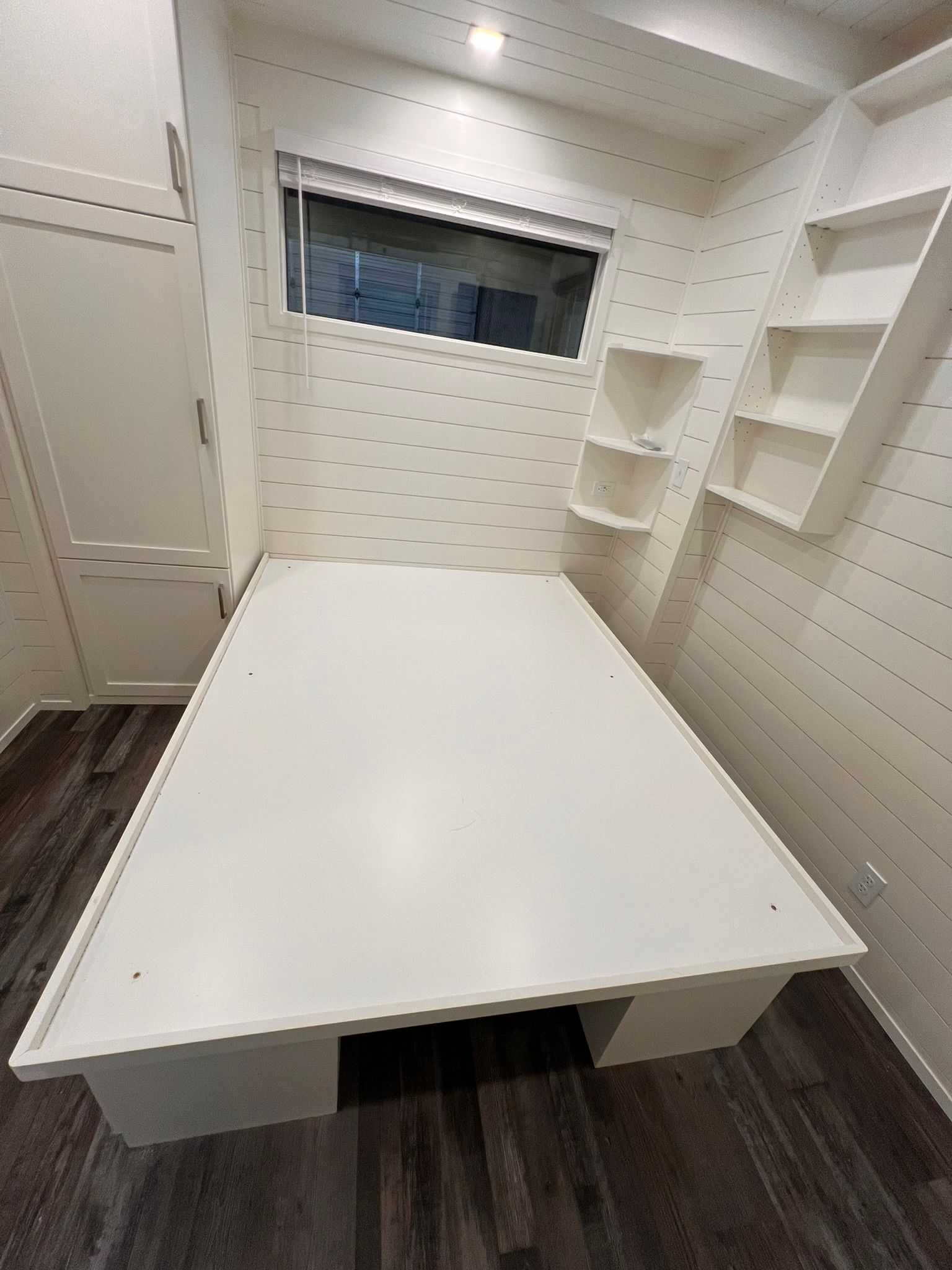 White-walled bedroom with a bed frame, shelves, window, and wood-look flooring.