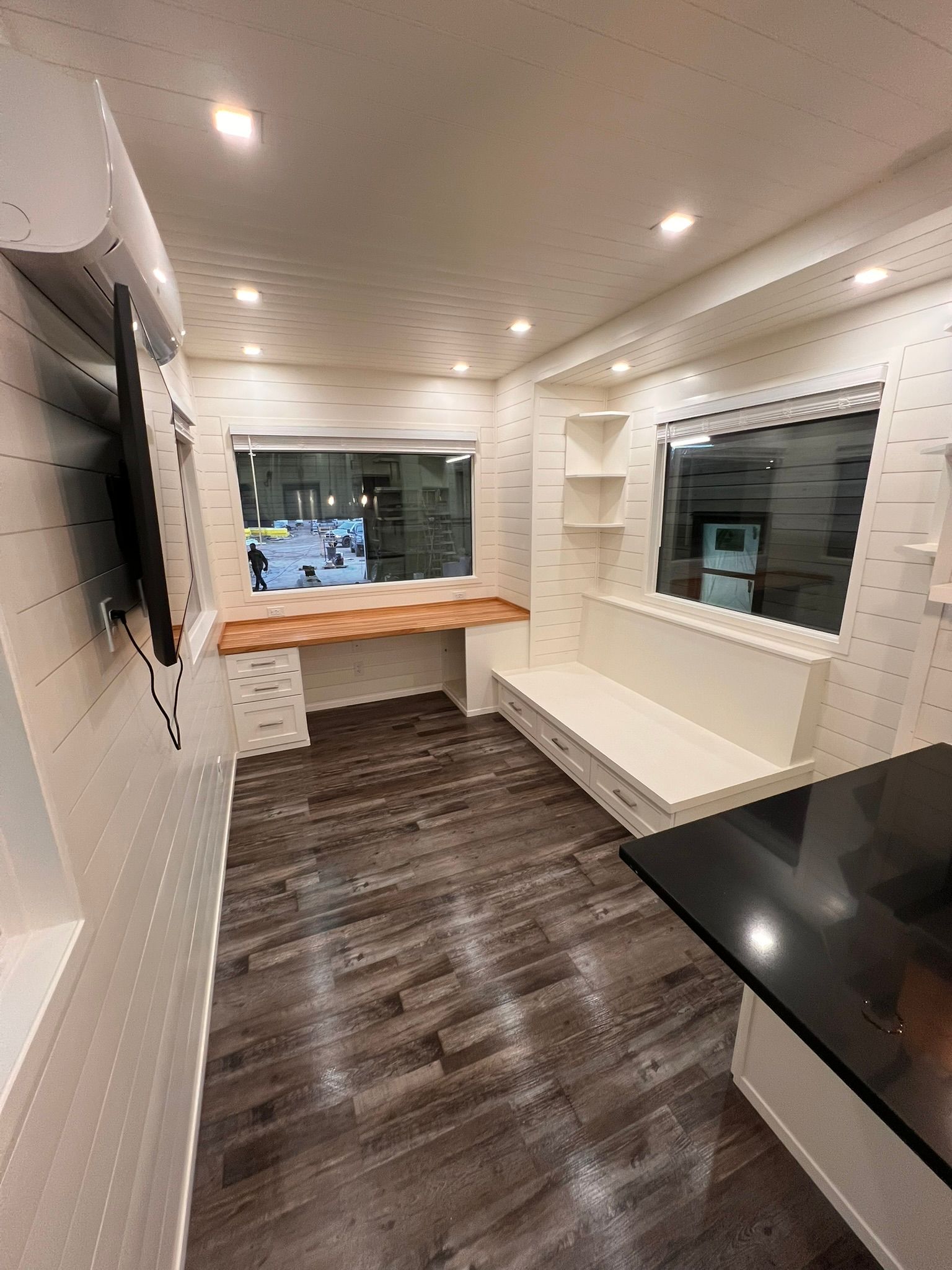 Interior of a tiny home with a wooden desk, built-in bench, and hardwood floors. Windows offer an outdoor view.