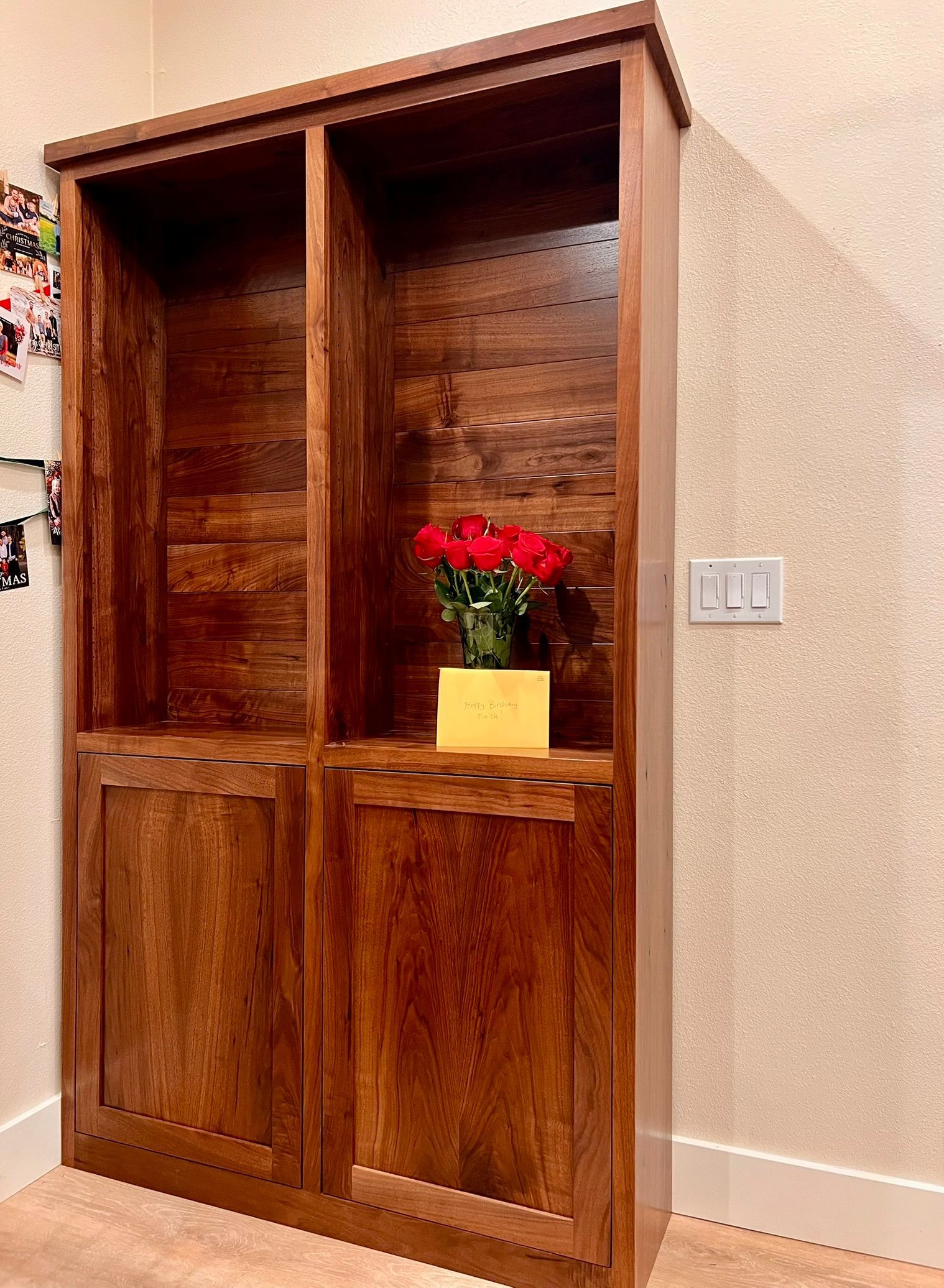 Wooden bookcase with two compartments, holding flowers. Brown wood, set against a wall.