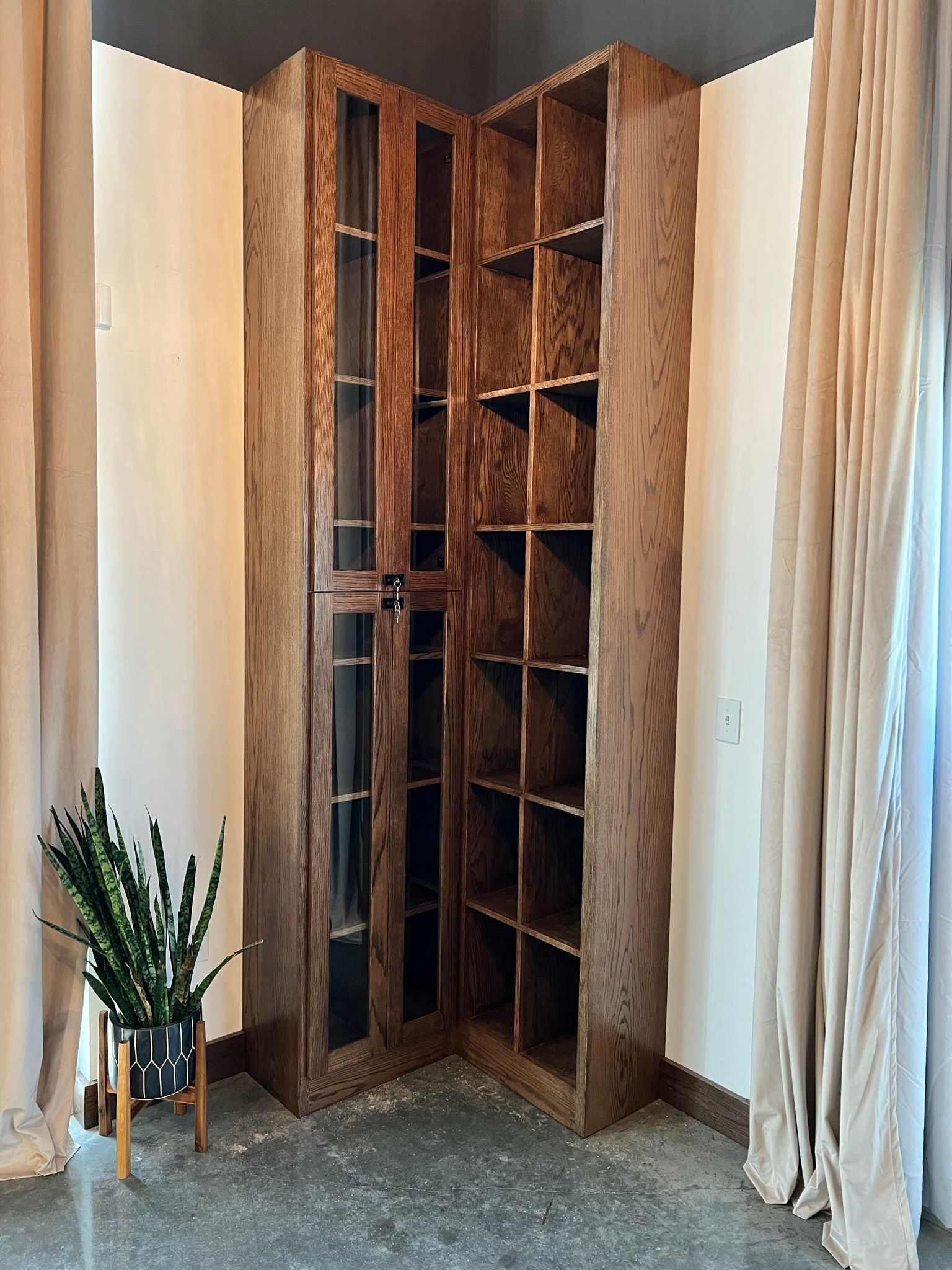 Corner wooden cabinet with shelves and glass doors, next to a potted plant and curtains.