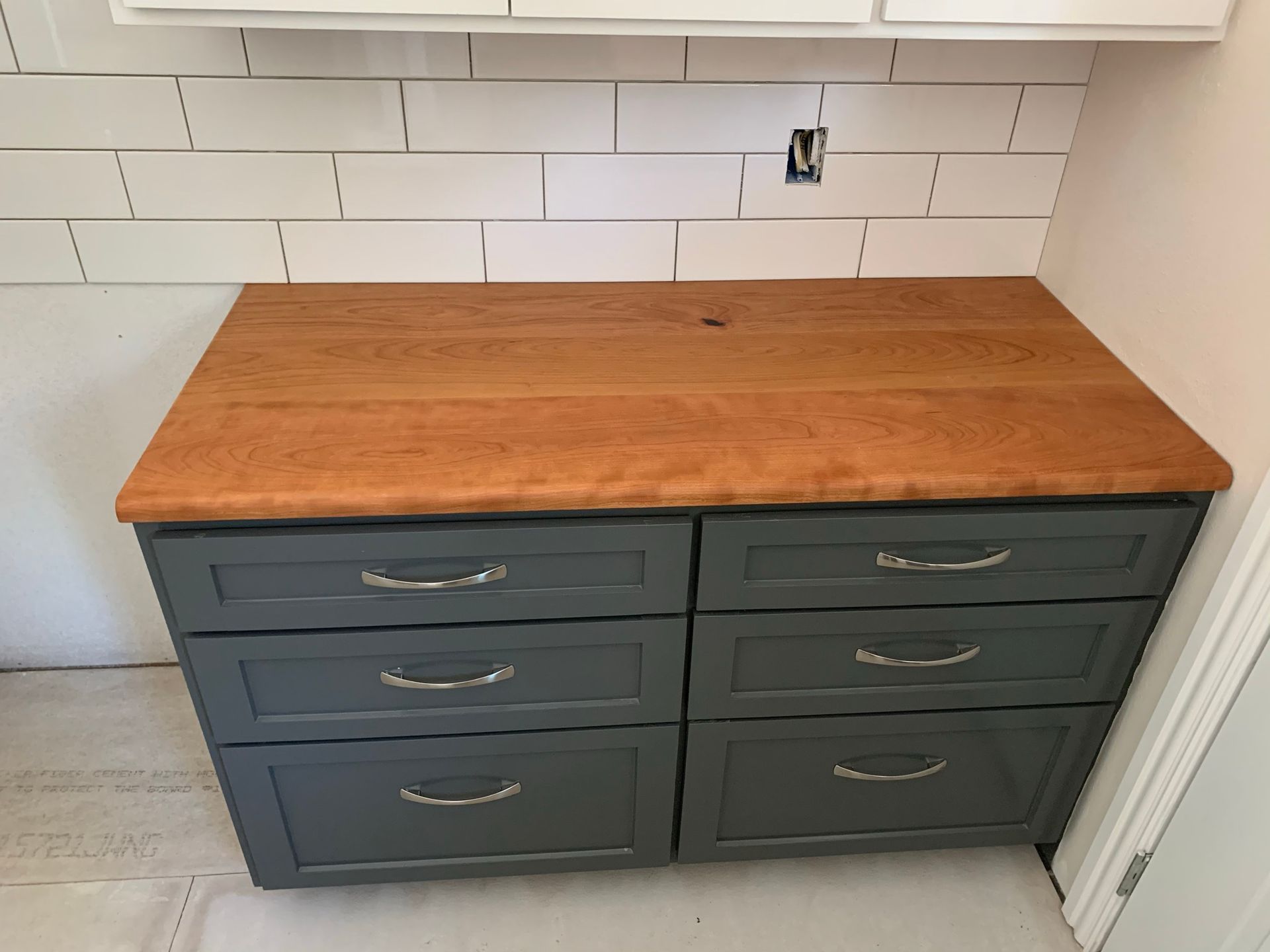 Gray cabinet with six drawers, wooden countertop, and white subway tile backsplash.
