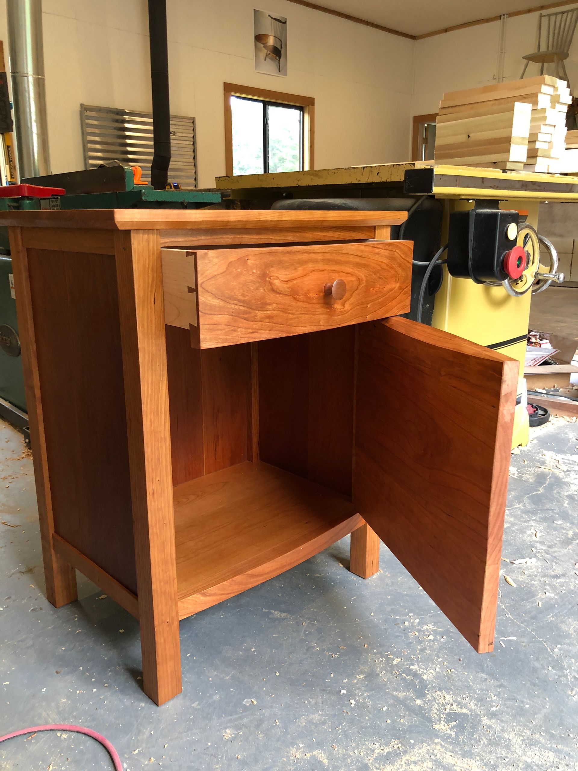 Wooden nightstand with open drawer and door in a workshop.