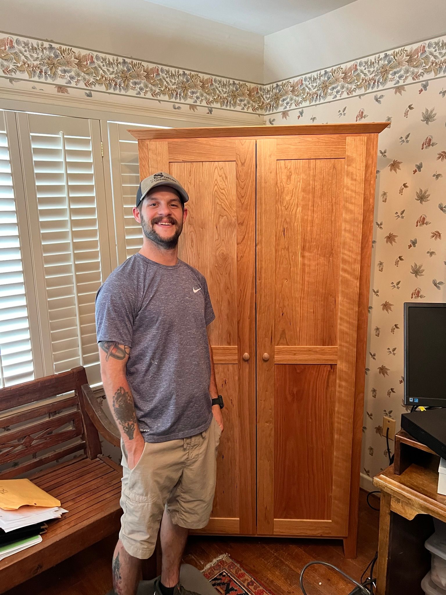 Man standing next to a large wooden cabinet in a room. Smiling, light-colored clothing.