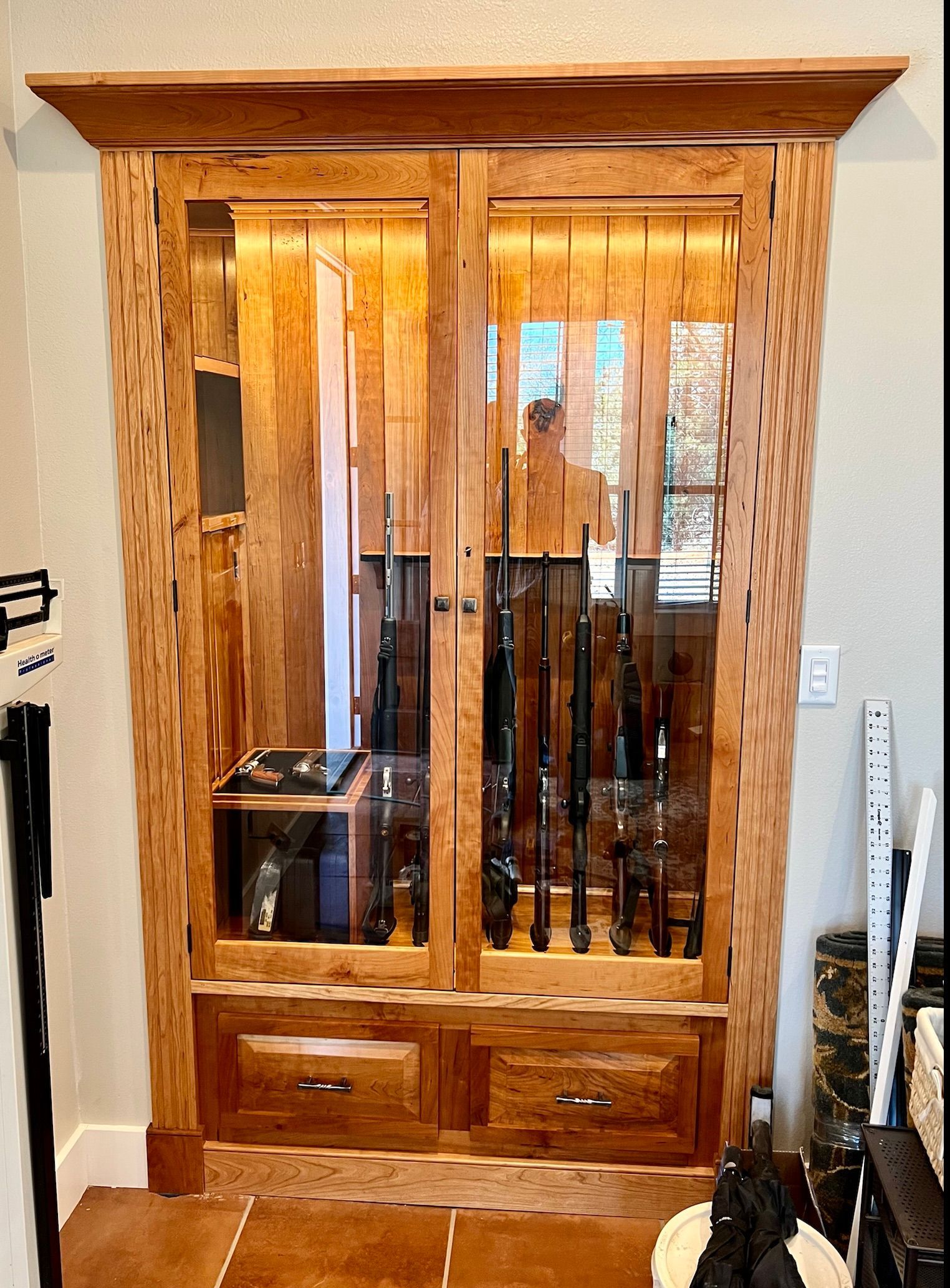 Wooden gun cabinet built into a wall with glass doors and drawers; guns inside.
