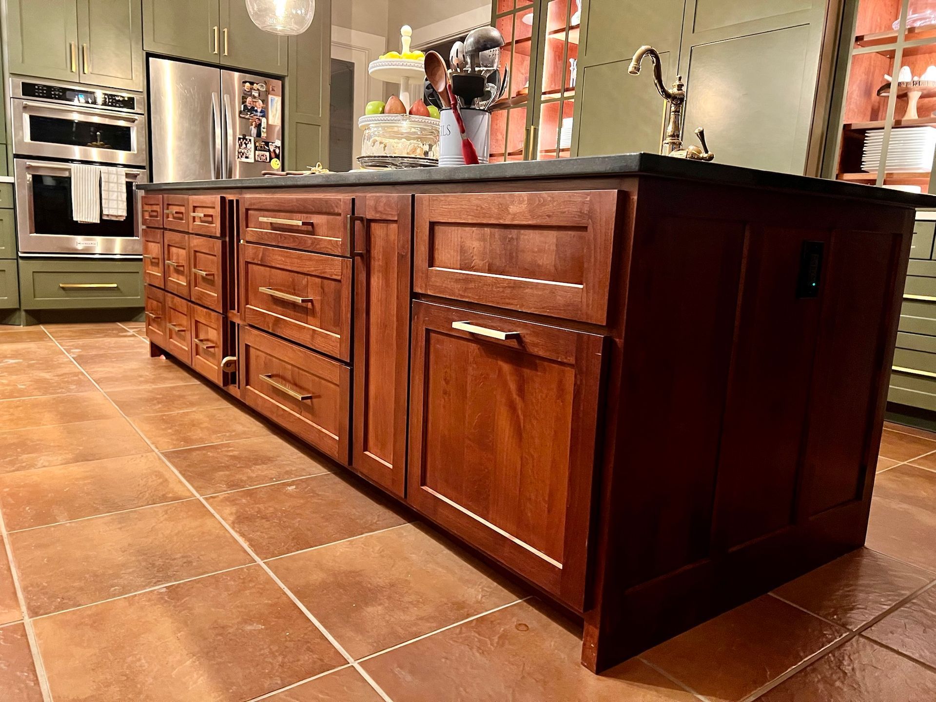 Brown kitchen island with dark countertop, multiple drawers and cabinets, on tile floor.