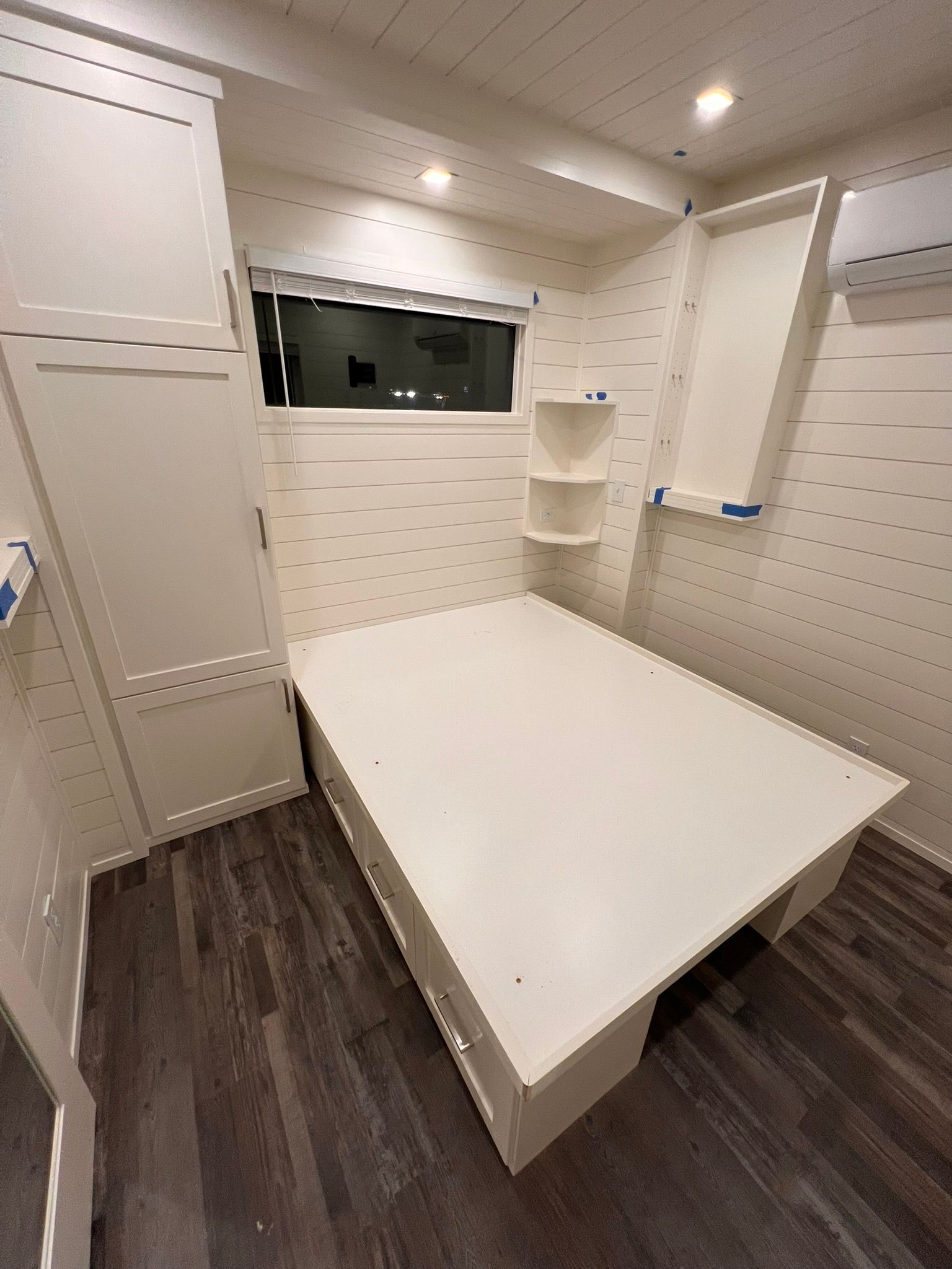 White built-in bed and storage in a small room with a window, shelves, and dark wood-look flooring.