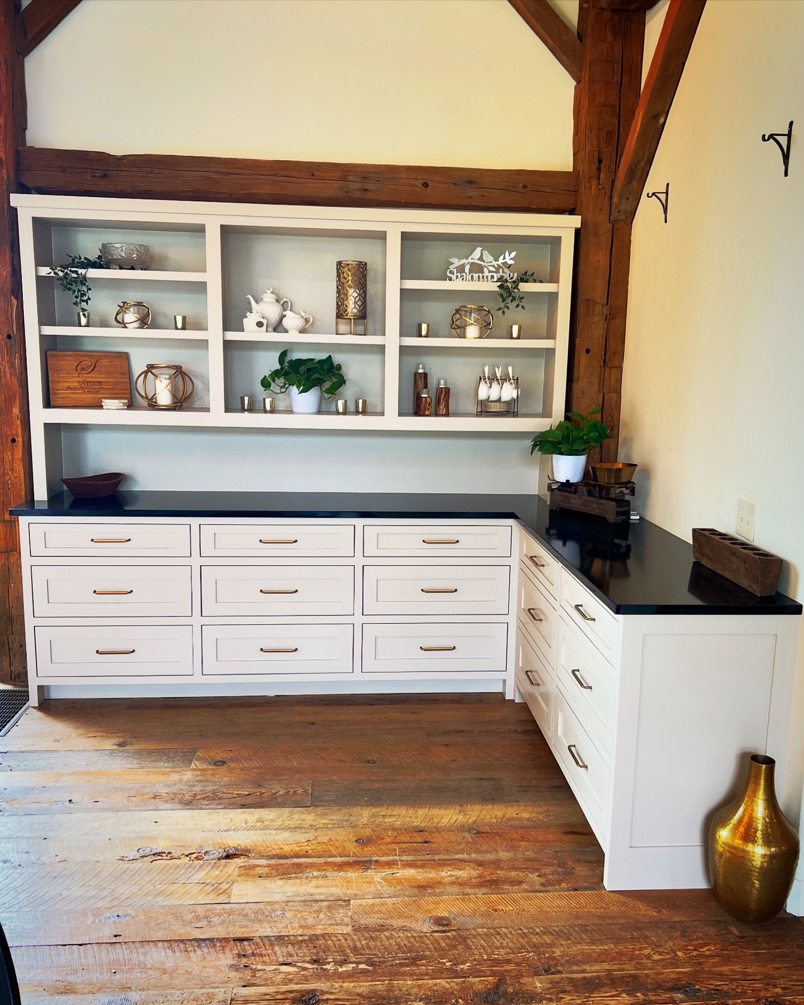 White cabinets with black countertops. Upper shelves display decor. Wooden floor and beam.