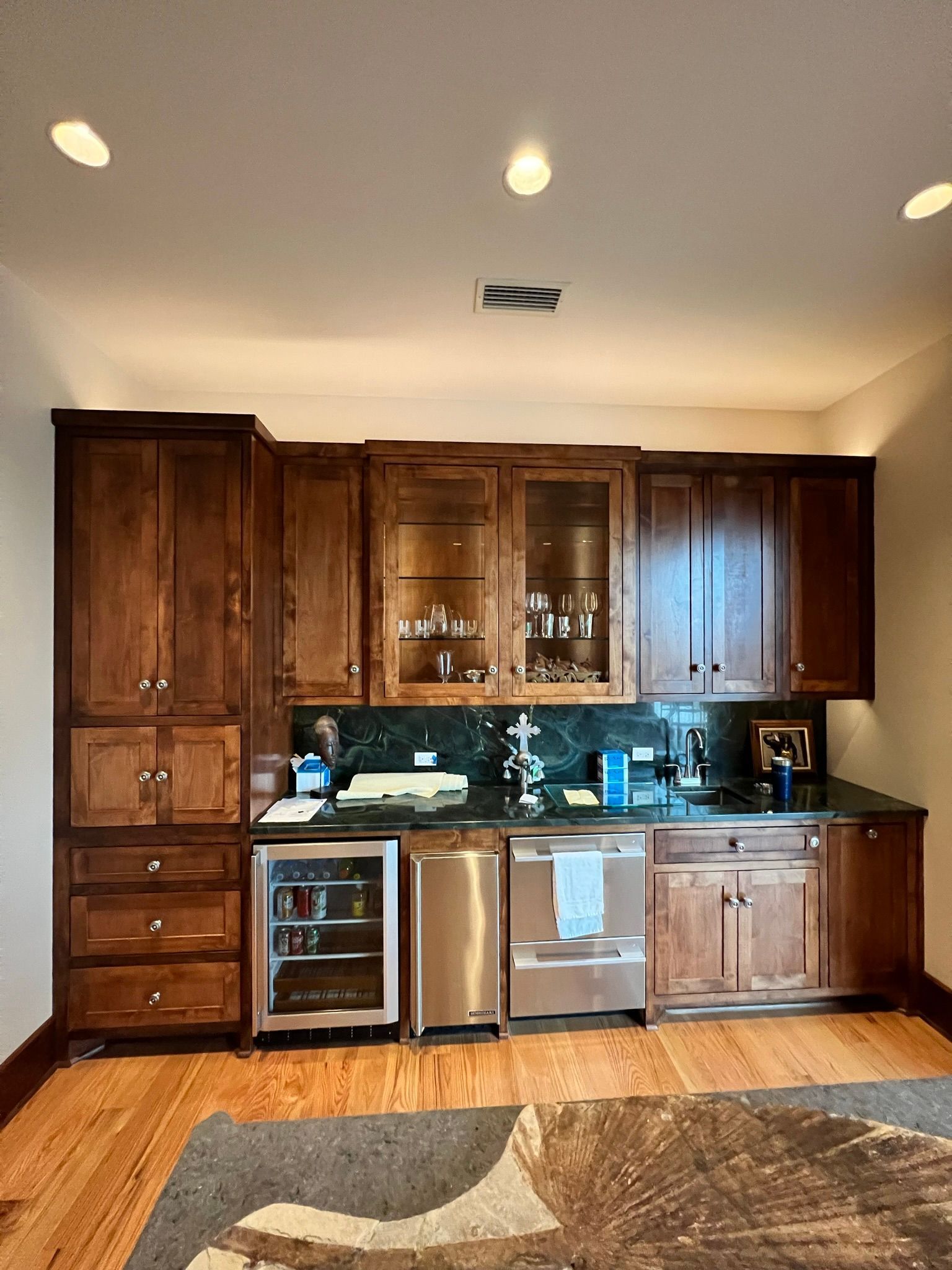 Wooden kitchen bar with cabinets, mini-fridge, dishwasher, sink, and countertop, with overhead lighting, on hardwood floor.