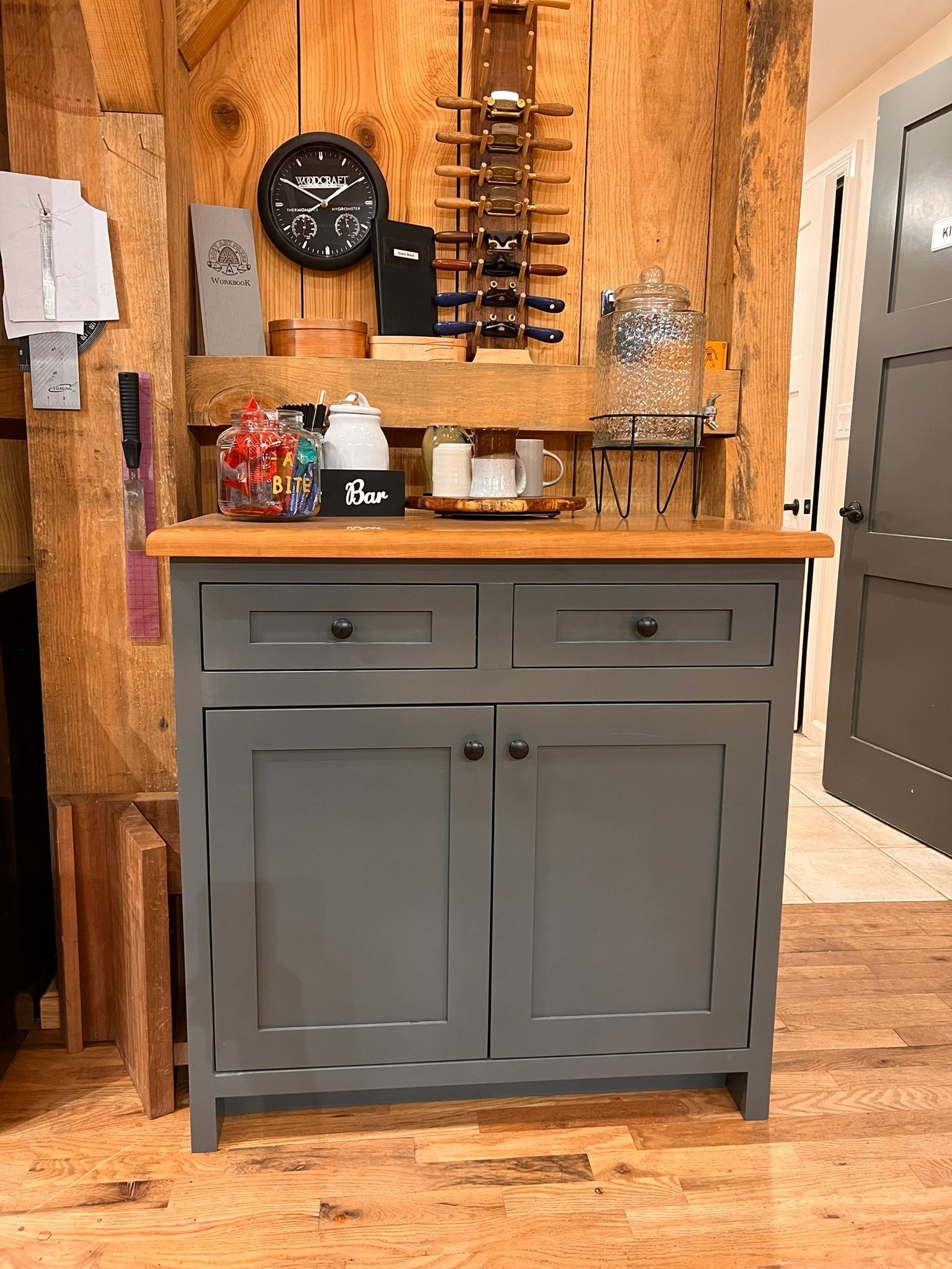 Gray cabinet with wooden countertop, holding a coffee station and wine rack, in a wood-paneled room.