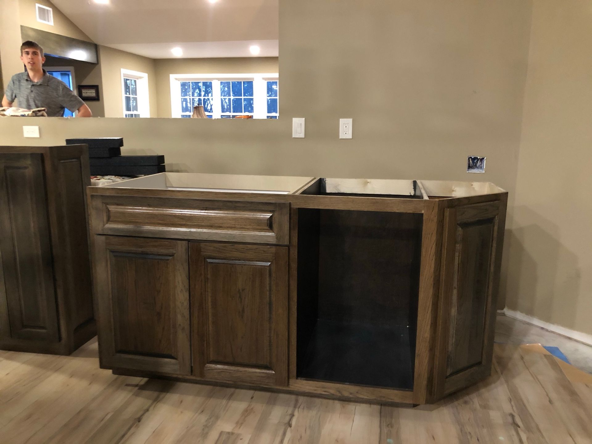 Kitchen cabinets, brown wood finish, under construction. Man in background, neutral beige walls, light flooring.