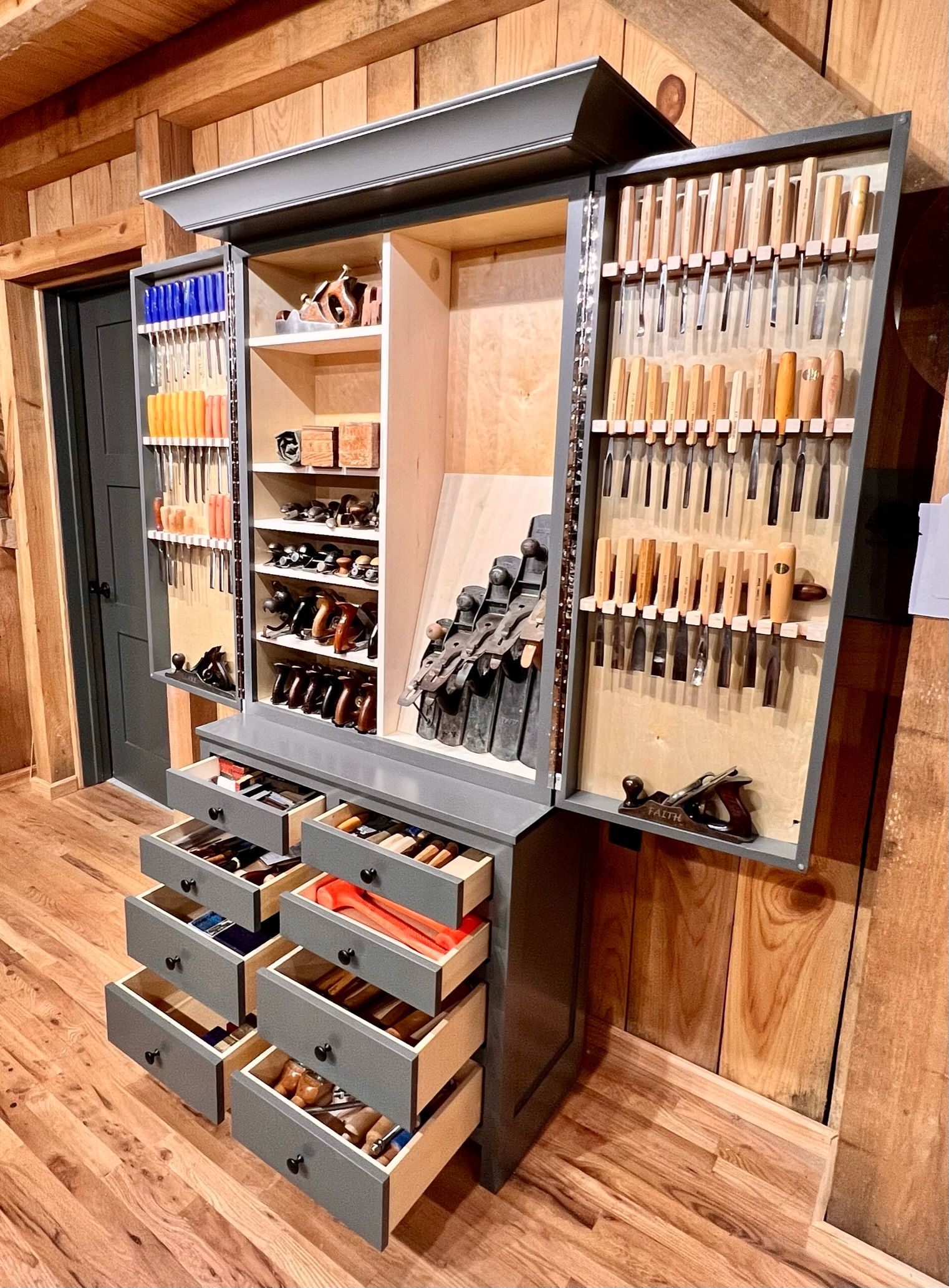 Gray wooden tool cabinet, drawers open, displaying carpentry tools in a wood-paneled workshop.