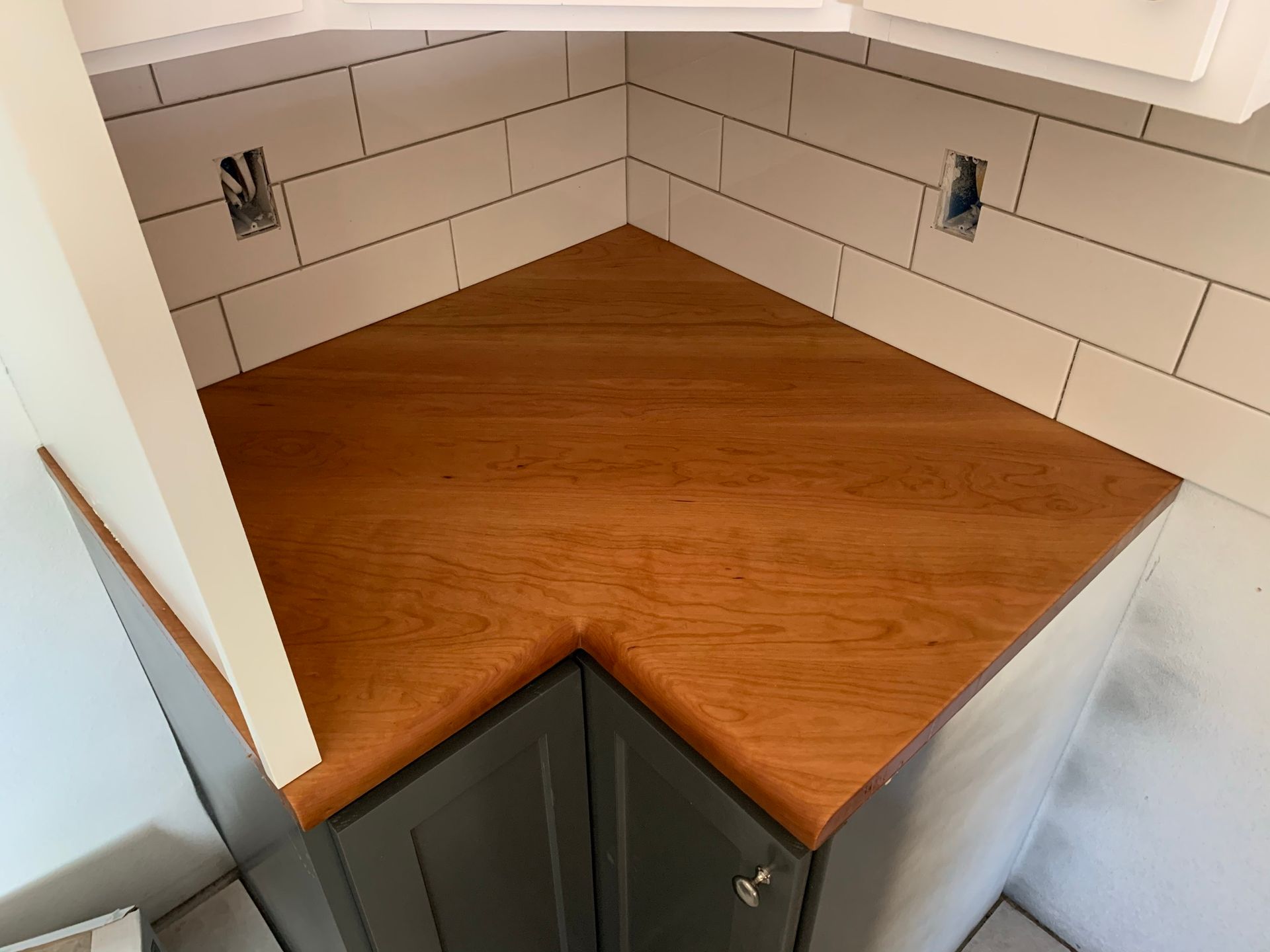 Wooden corner countertop over gray cabinets, with white tiled backsplash.