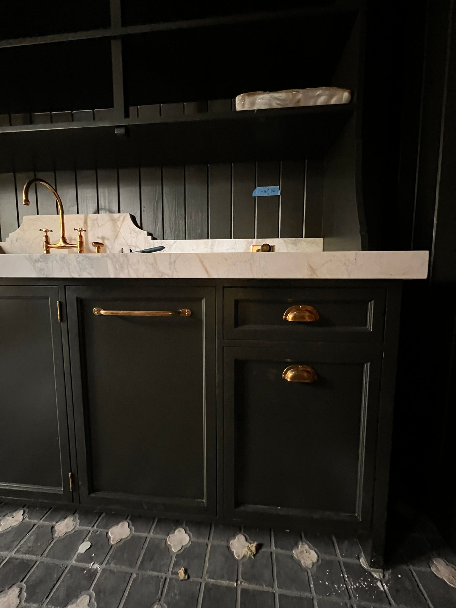 Dark-painted cabinet with gold hardware, marble countertop, and shelves. Sink with gold faucet. Black and gray tiled floor.