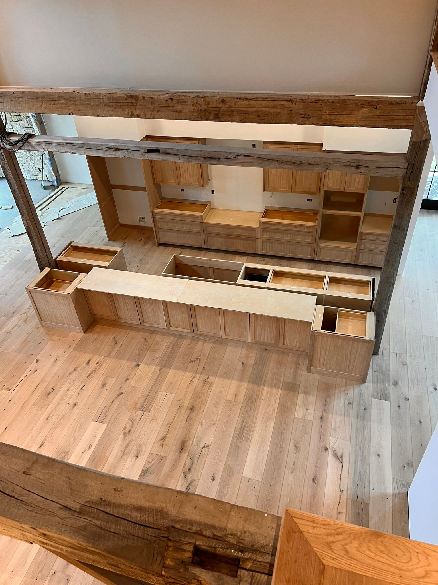 Wooden kitchen cabinetry and island, unfinished, in a light-floored room, with exposed beams overhead.