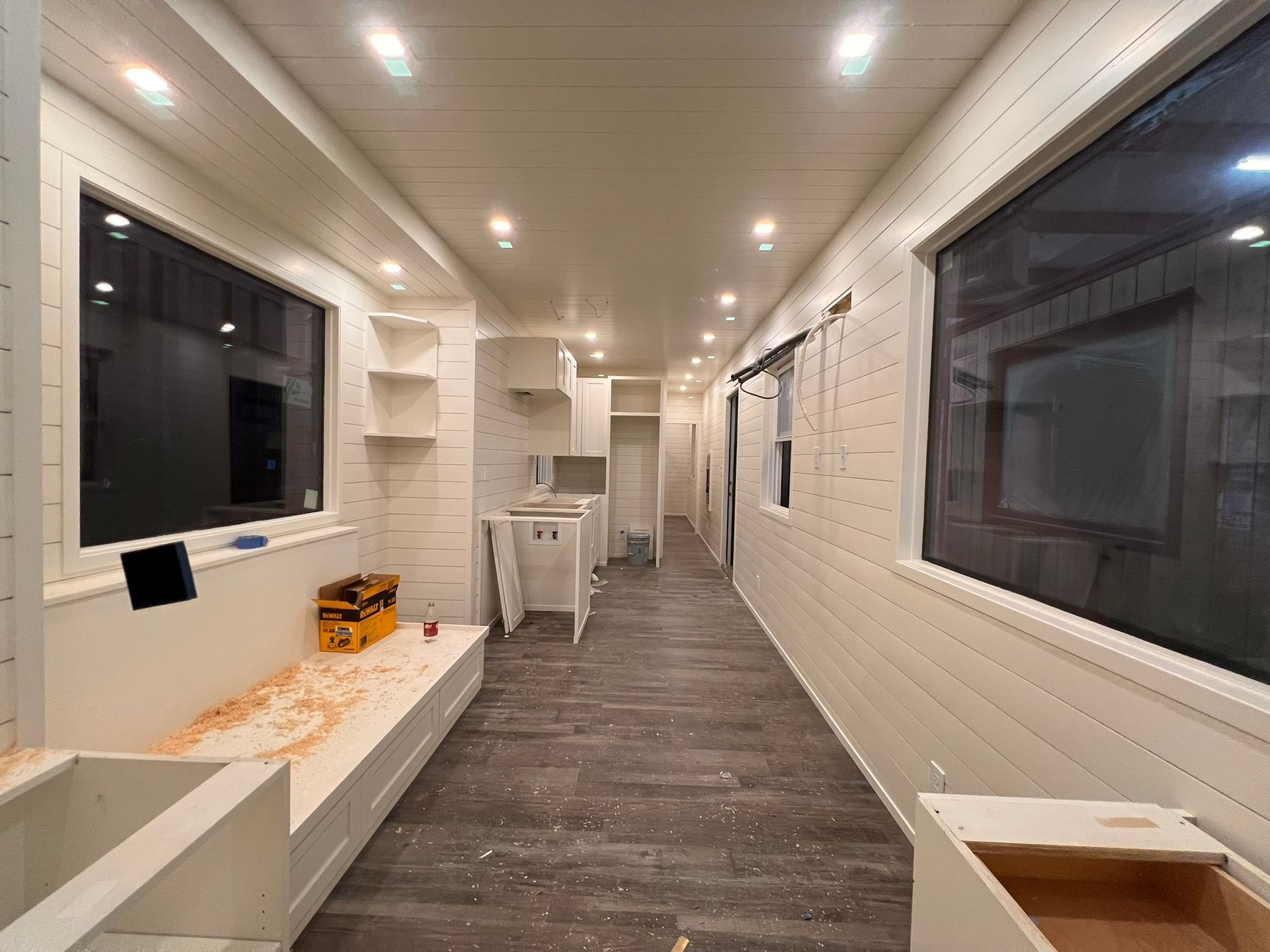 Interior of a narrow, white-walled room with wood flooring, windows, and shelving.