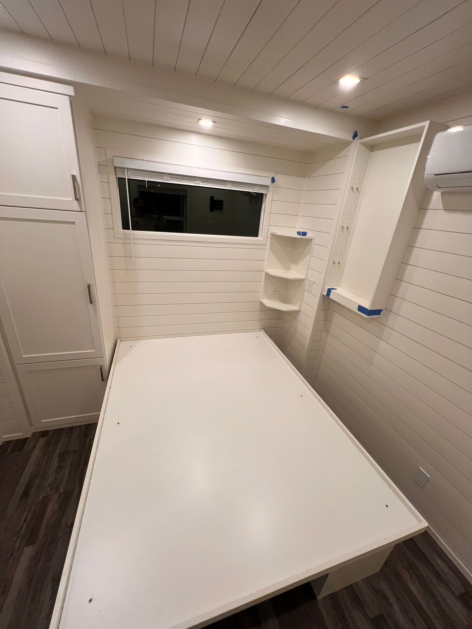 Interior of a camper van with a white fold-down bed, small shelves, and a window.