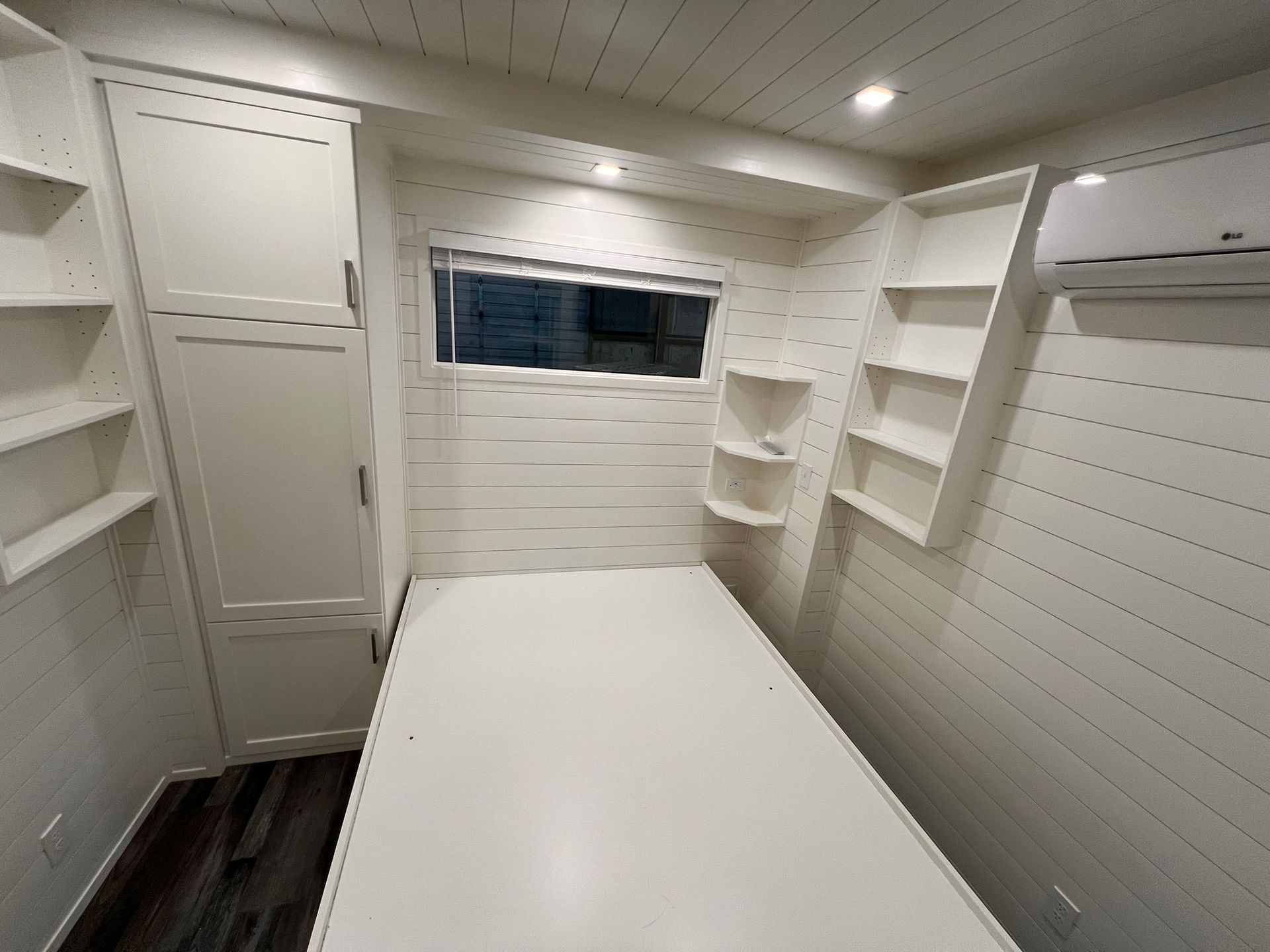 Small white bedroom with bed, shelves, window, and air conditioner.