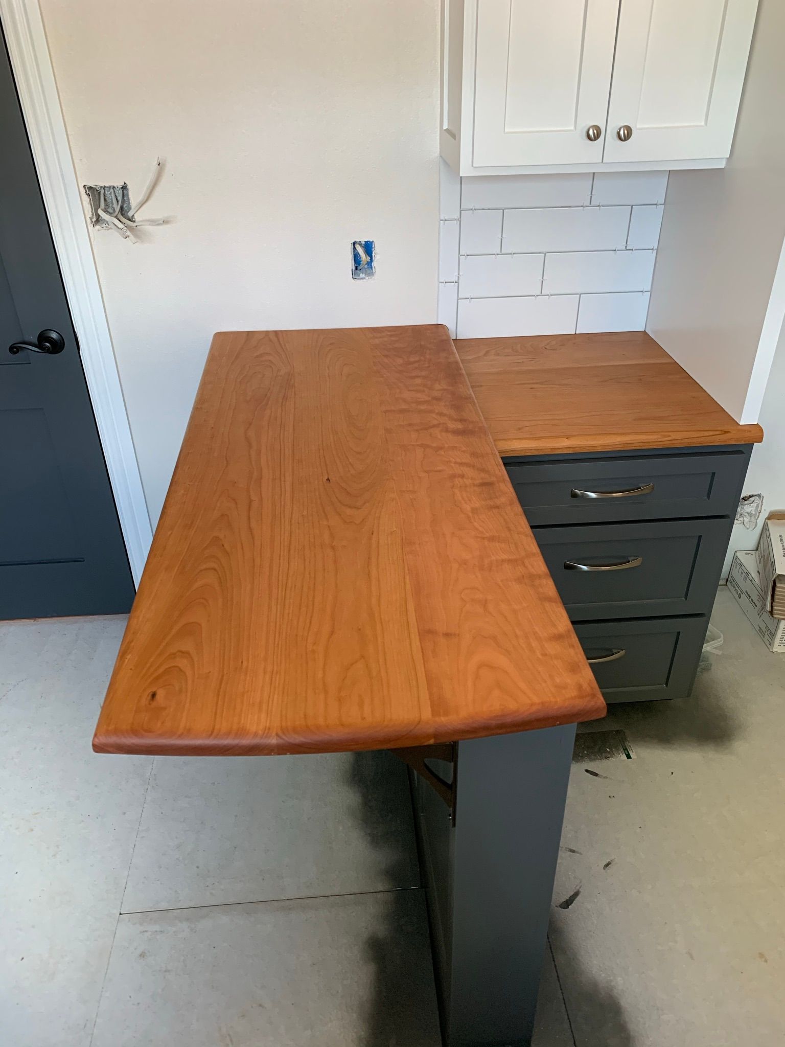 L-shaped wooden countertop on grey cabinets in a kitchen under construction. White cabinets are visible.
