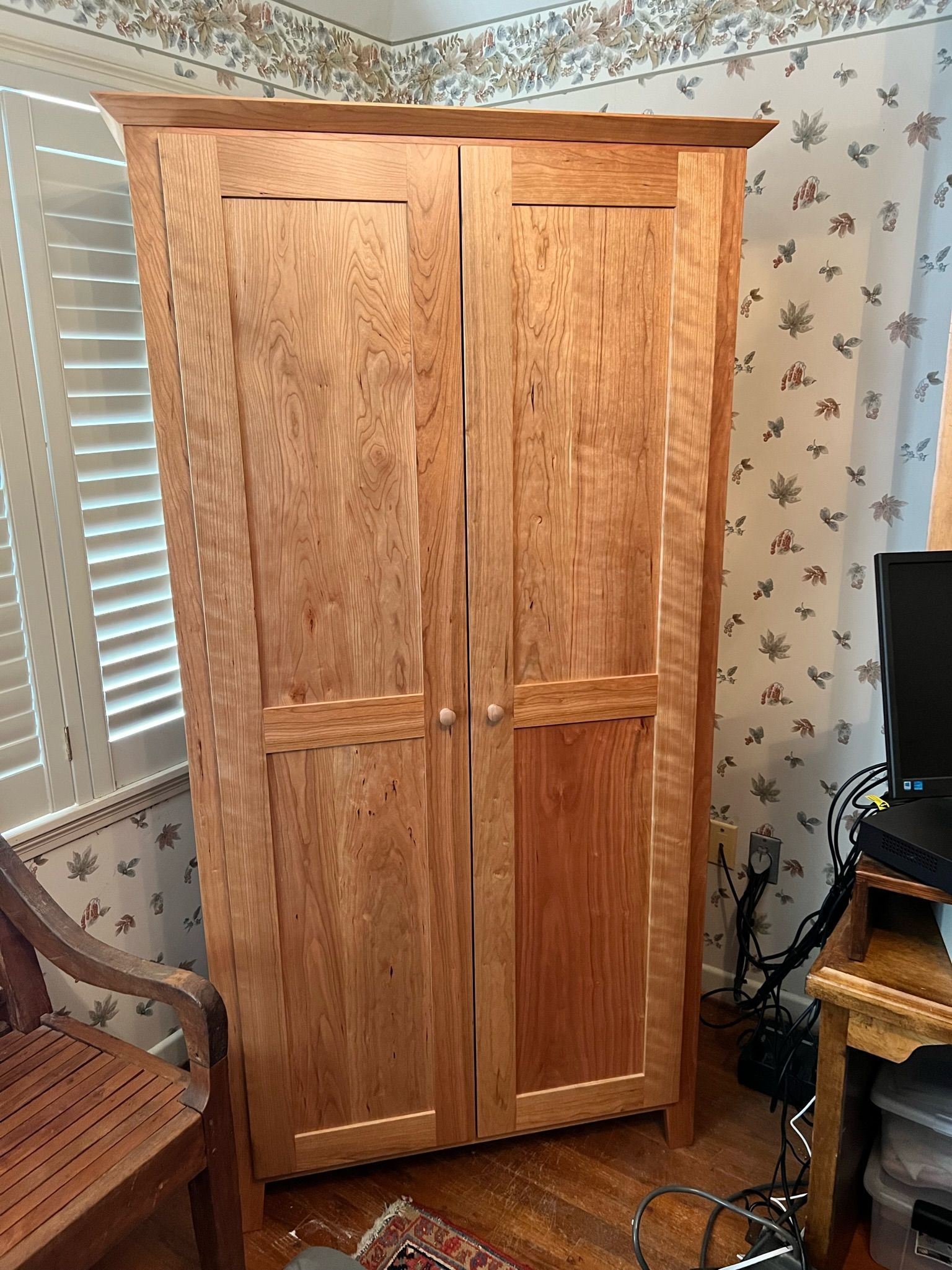 Tall wooden cabinet with two doors in a room, next to a chair and a desk.