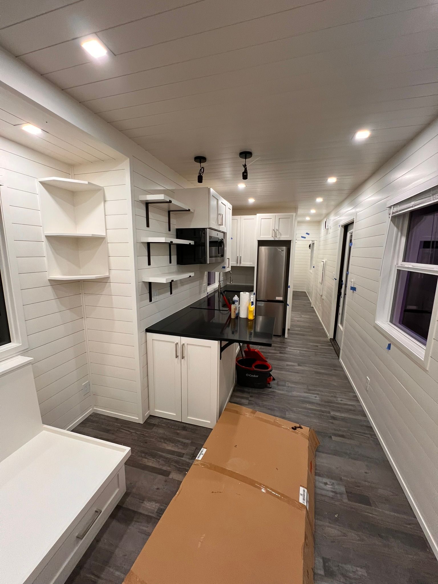 Long, narrow kitchen with white cabinets, black countertops, and dark floor; cardboard box in the foreground.