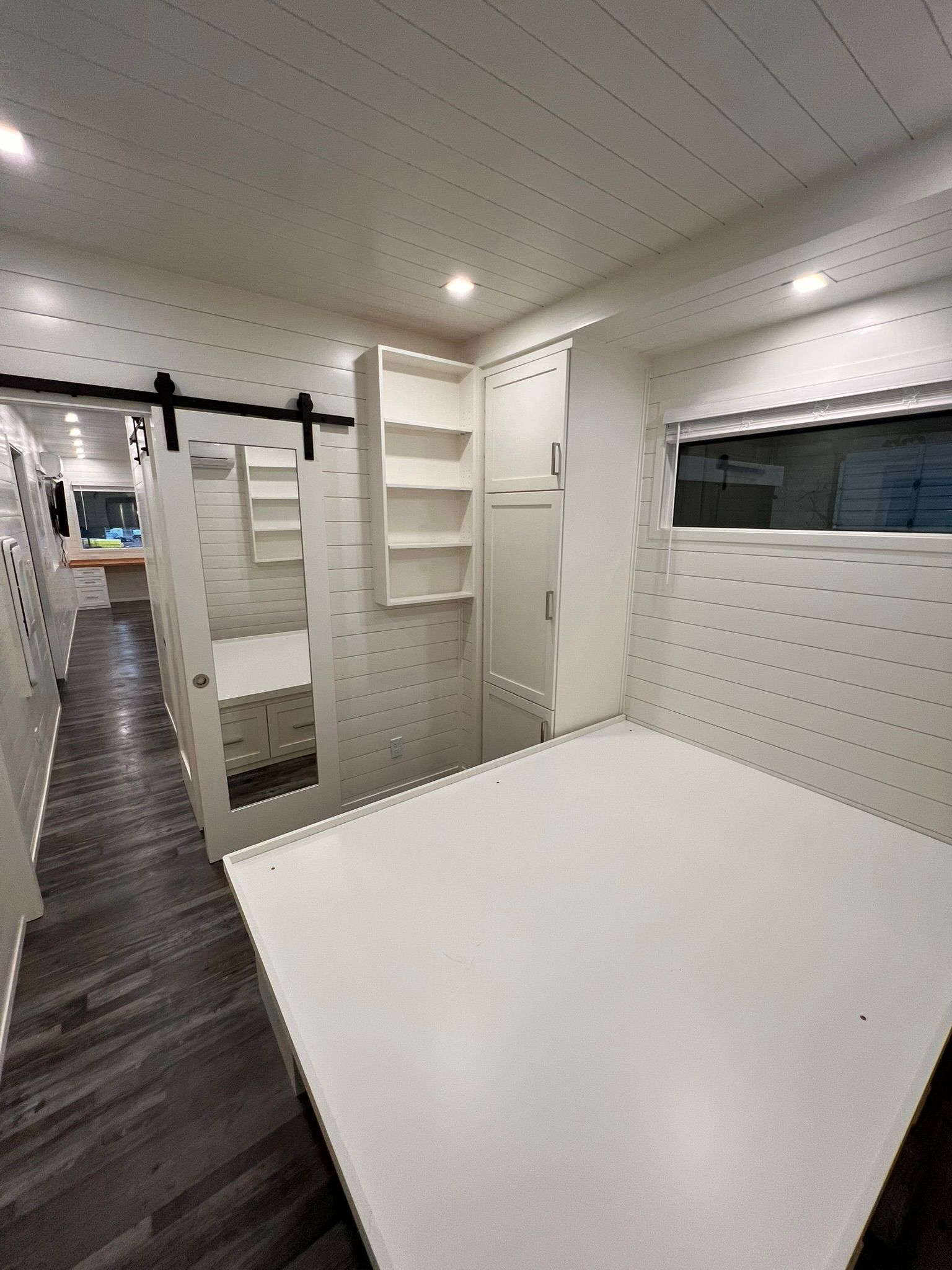 Interior view of a white mobile home. Features include a bed, storage, and dark floorboards.