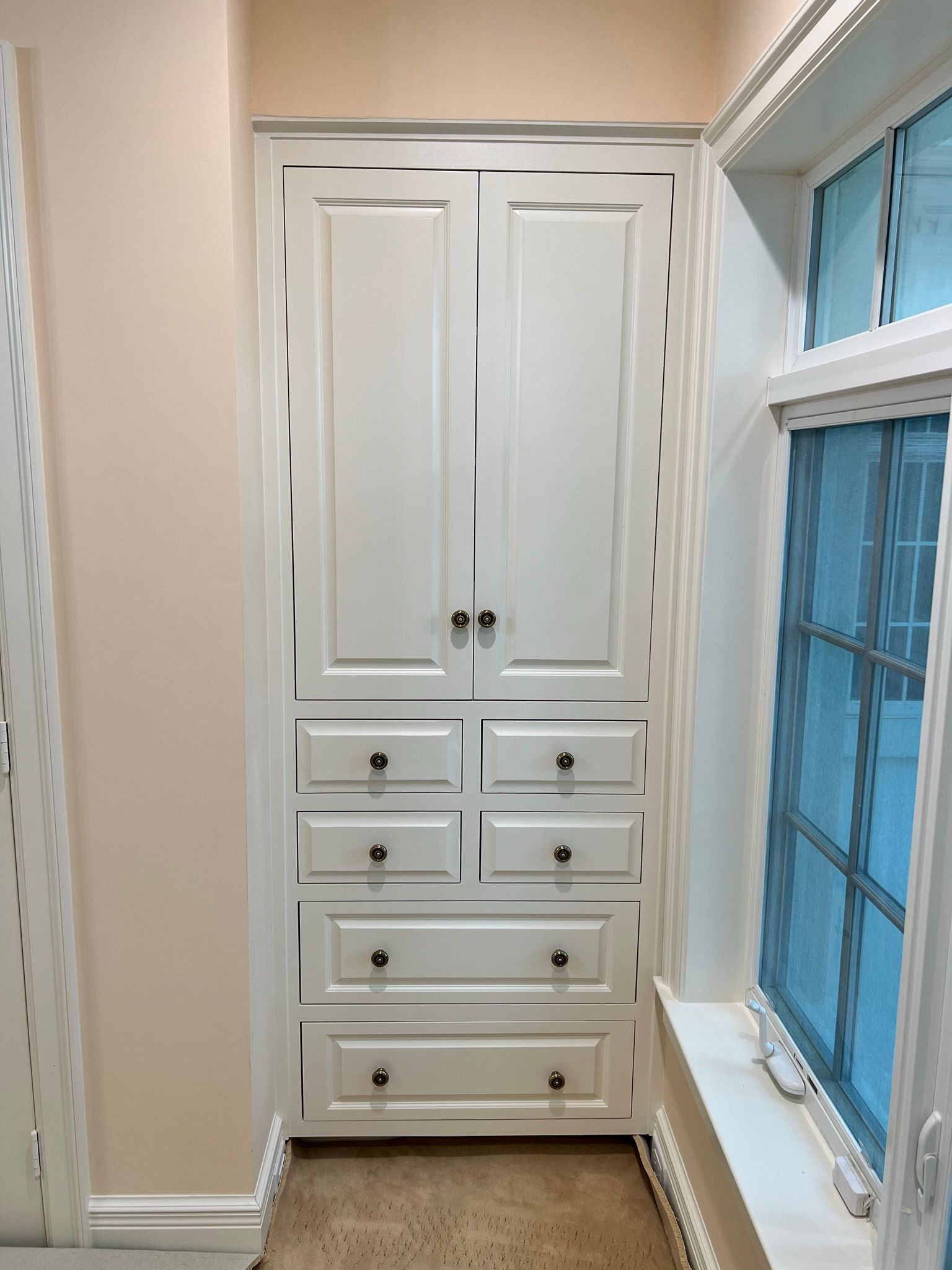 White built-in cabinet with two doors and drawers, next to a window with blue tint.