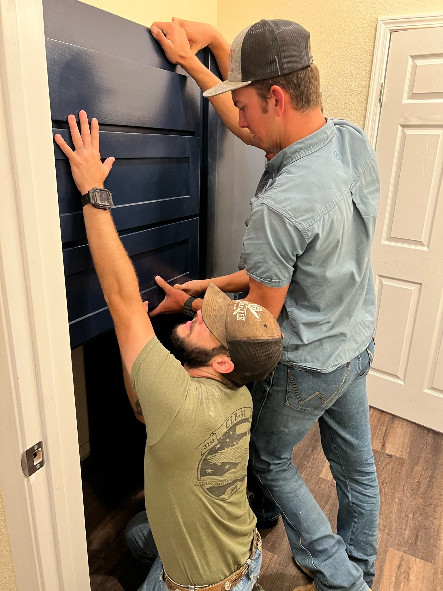 Two men installing a blue cabinet in a room with a white door. 