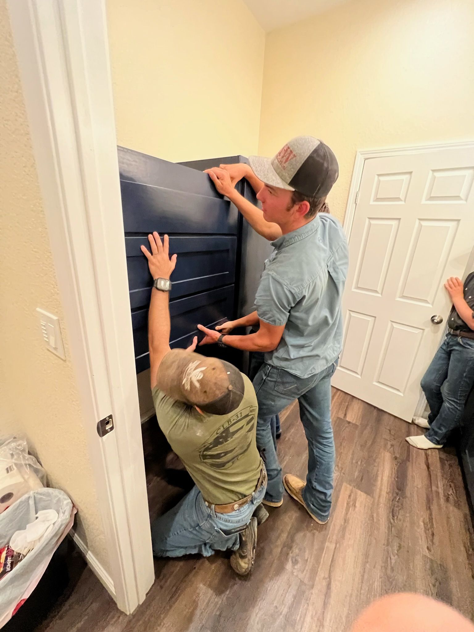 Two people moving a large blue object into a doorway. One kneels, the other stands. White door and beige wall.