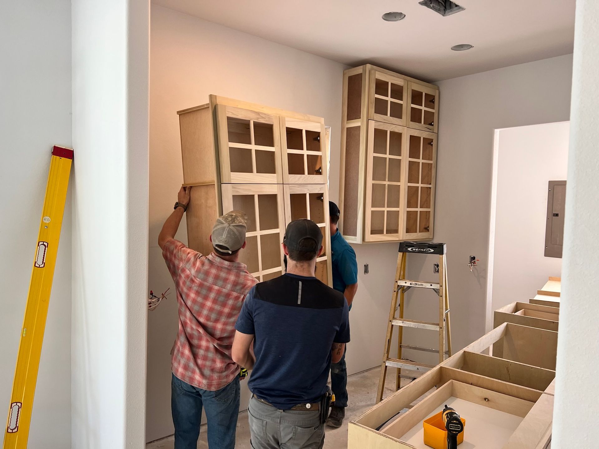 Three workers installing wooden kitchen cabinets on a white wall; tools and ladder present.