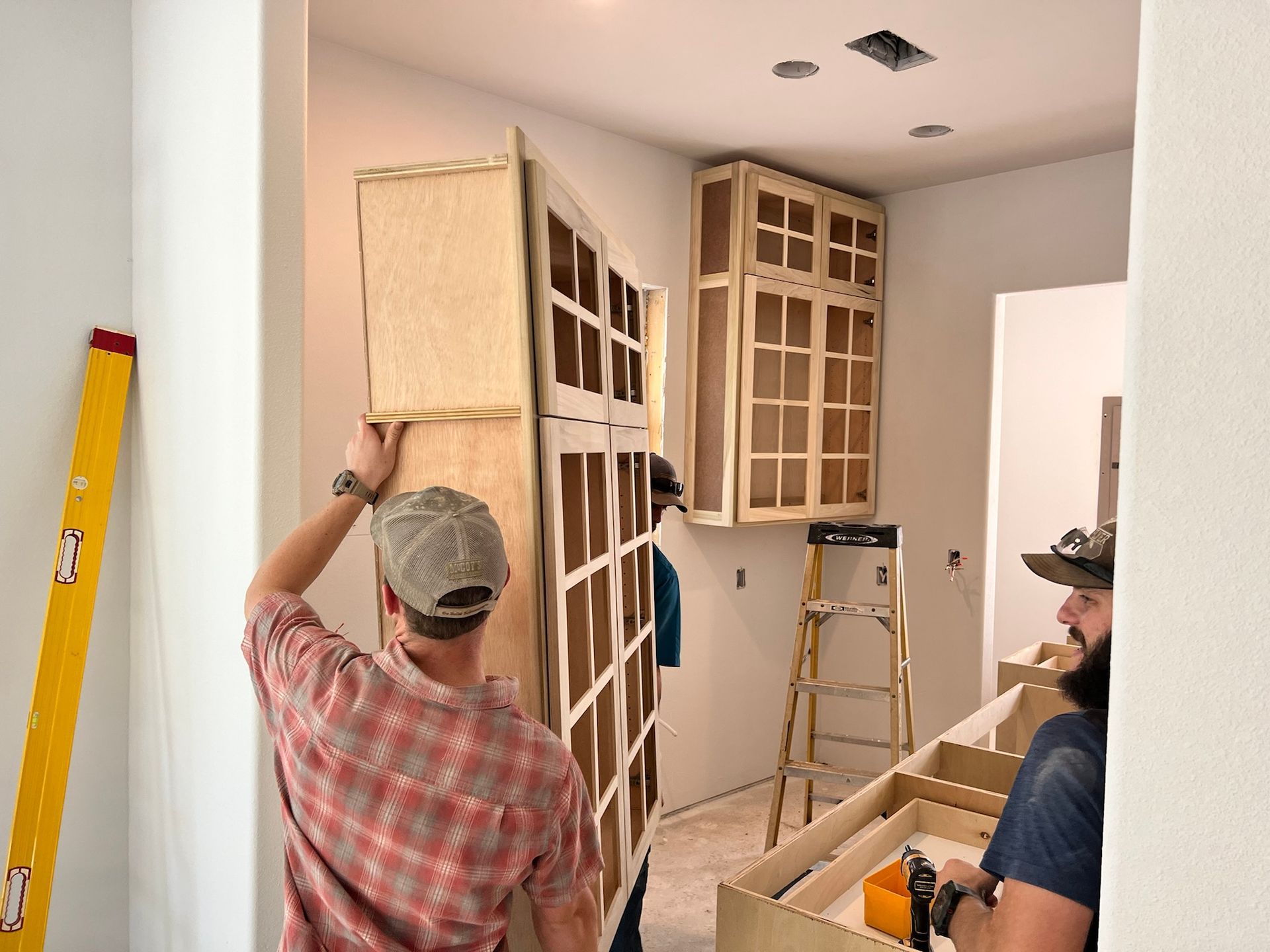 Two people installing unfinished kitchen cabinets. One holds a cabinet, the other works on base cabinets near a ladder.