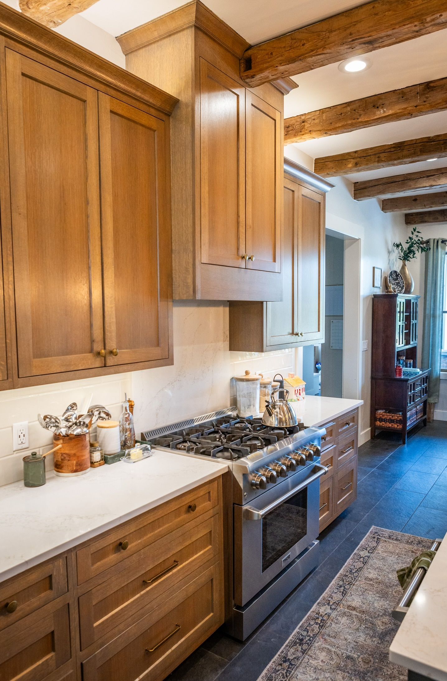 Wooden kitchen cabinets, stainless steel range, white countertops, exposed ceiling beams, and a dark wooden floor.