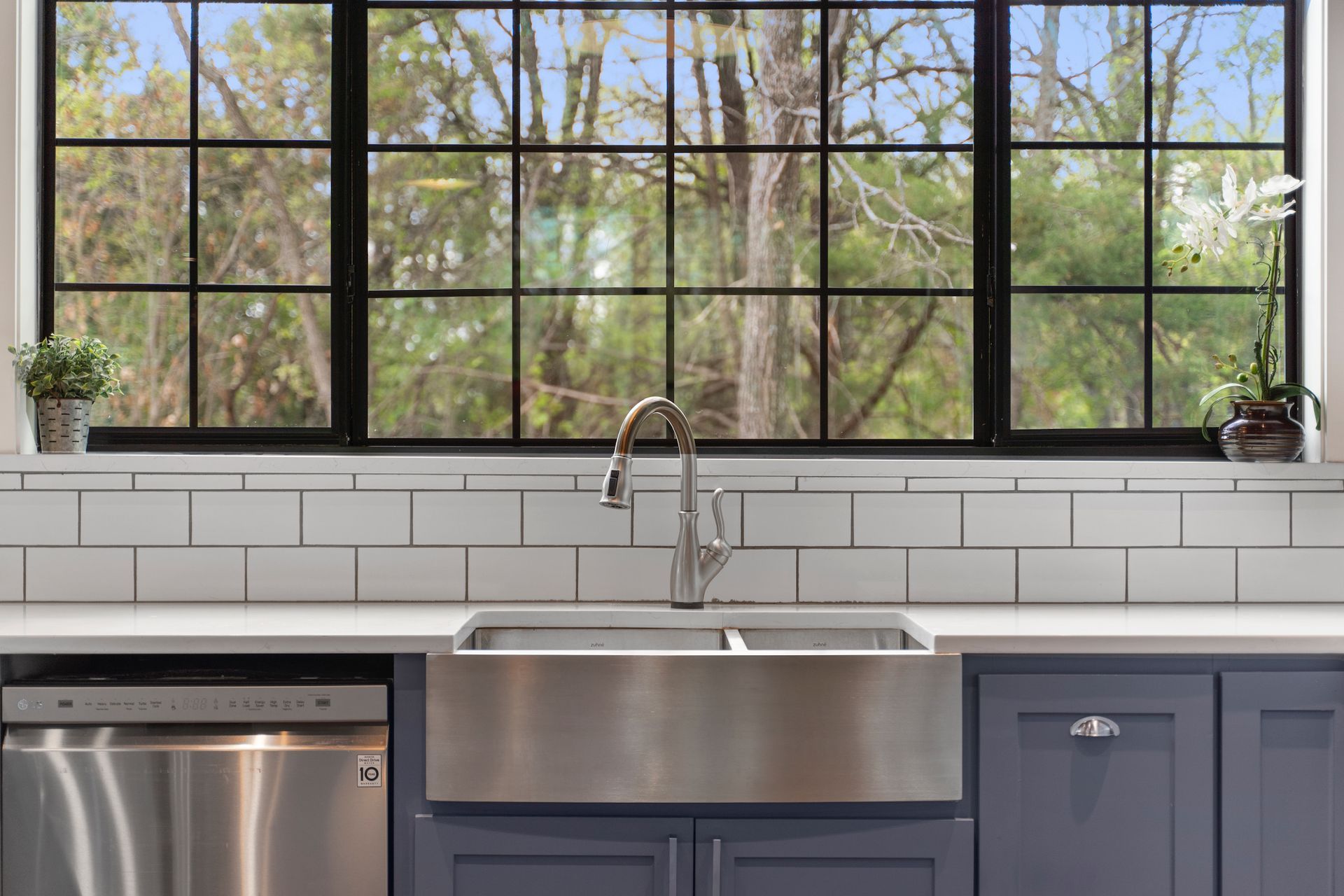 Kitchen sink with stainless steel basin, faucet, and dishwasher, beneath a window with a view of trees.