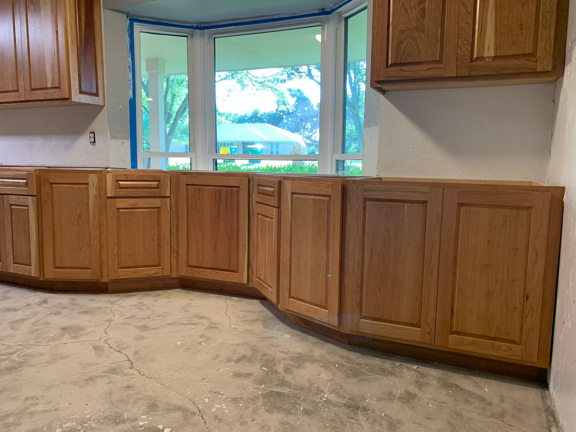 Kitchen cabinets, light brown wood, installed against a wall with a window. Concrete floor.
