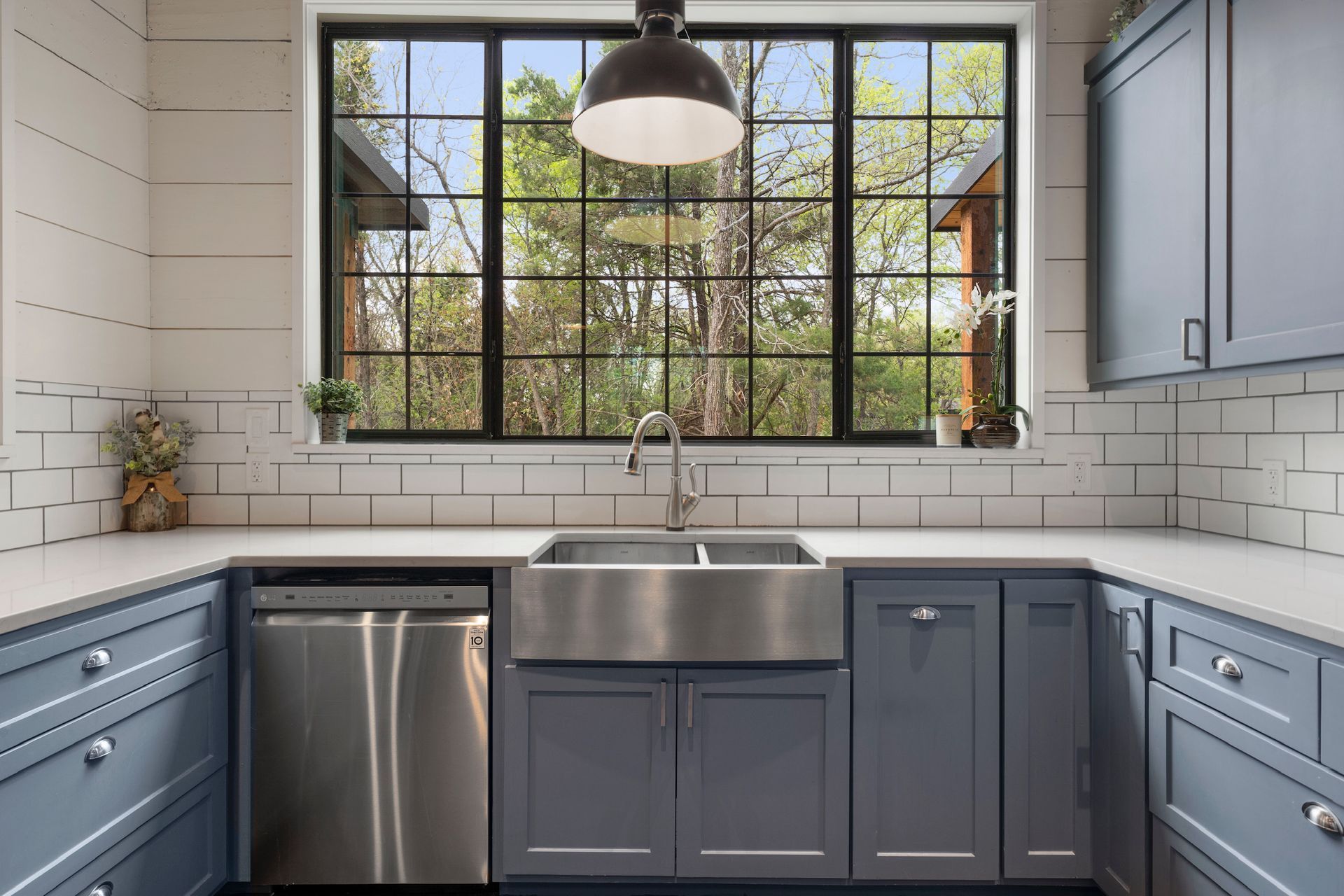 Blue kitchen with stainless steel sink, cabinets, dishwasher, and large window overlooking trees.