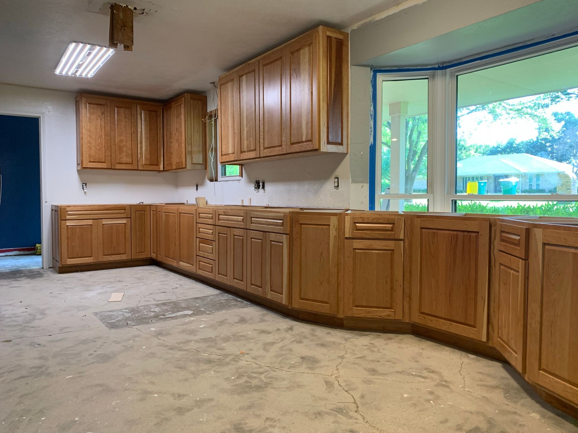 Kitchen renovation: light wood cabinets installed, unfinished concrete floor, large bay window, blue wall.