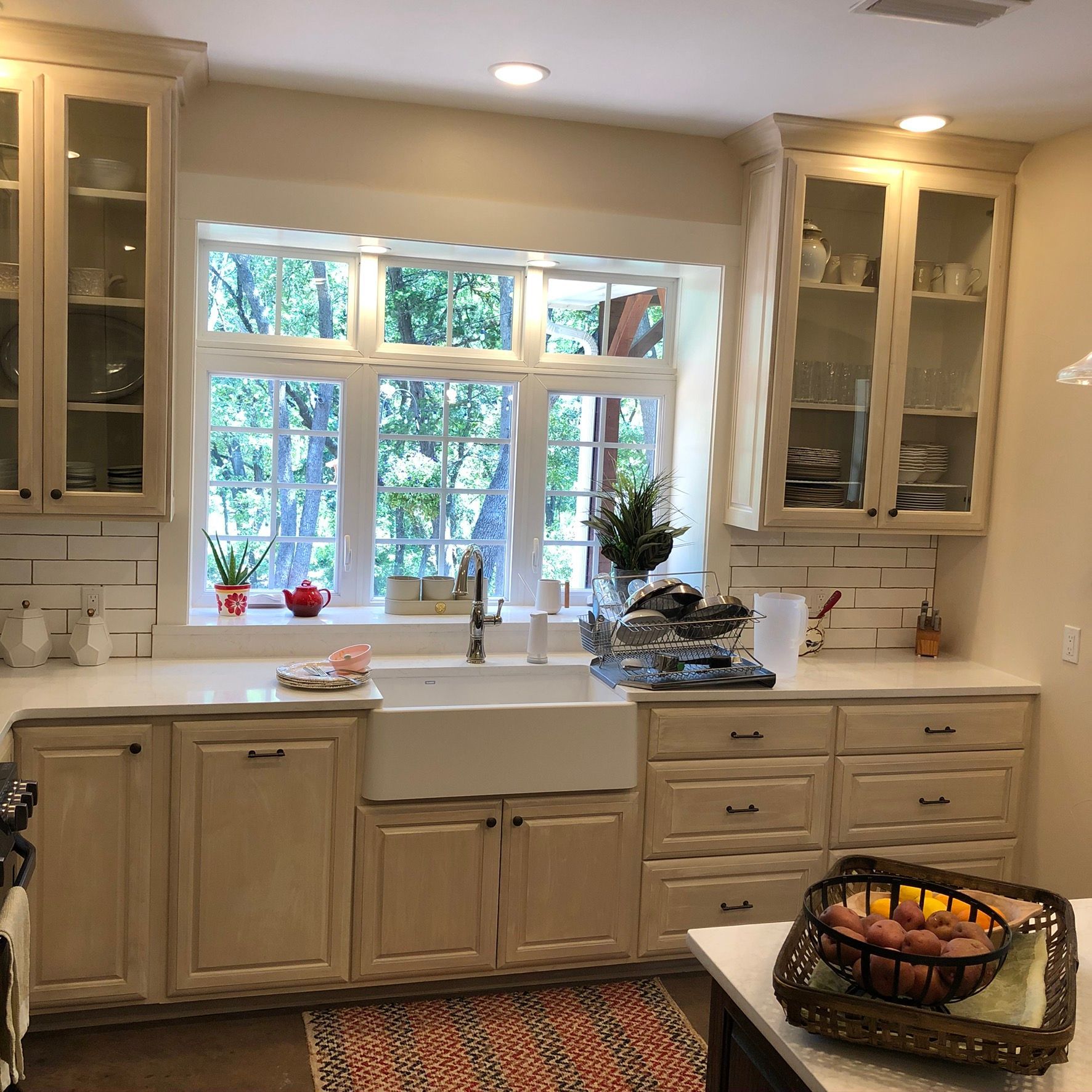 Beige kitchen with farmhouse sink, window overlooking trees, cabinets and drawers, fruit basket on counter.