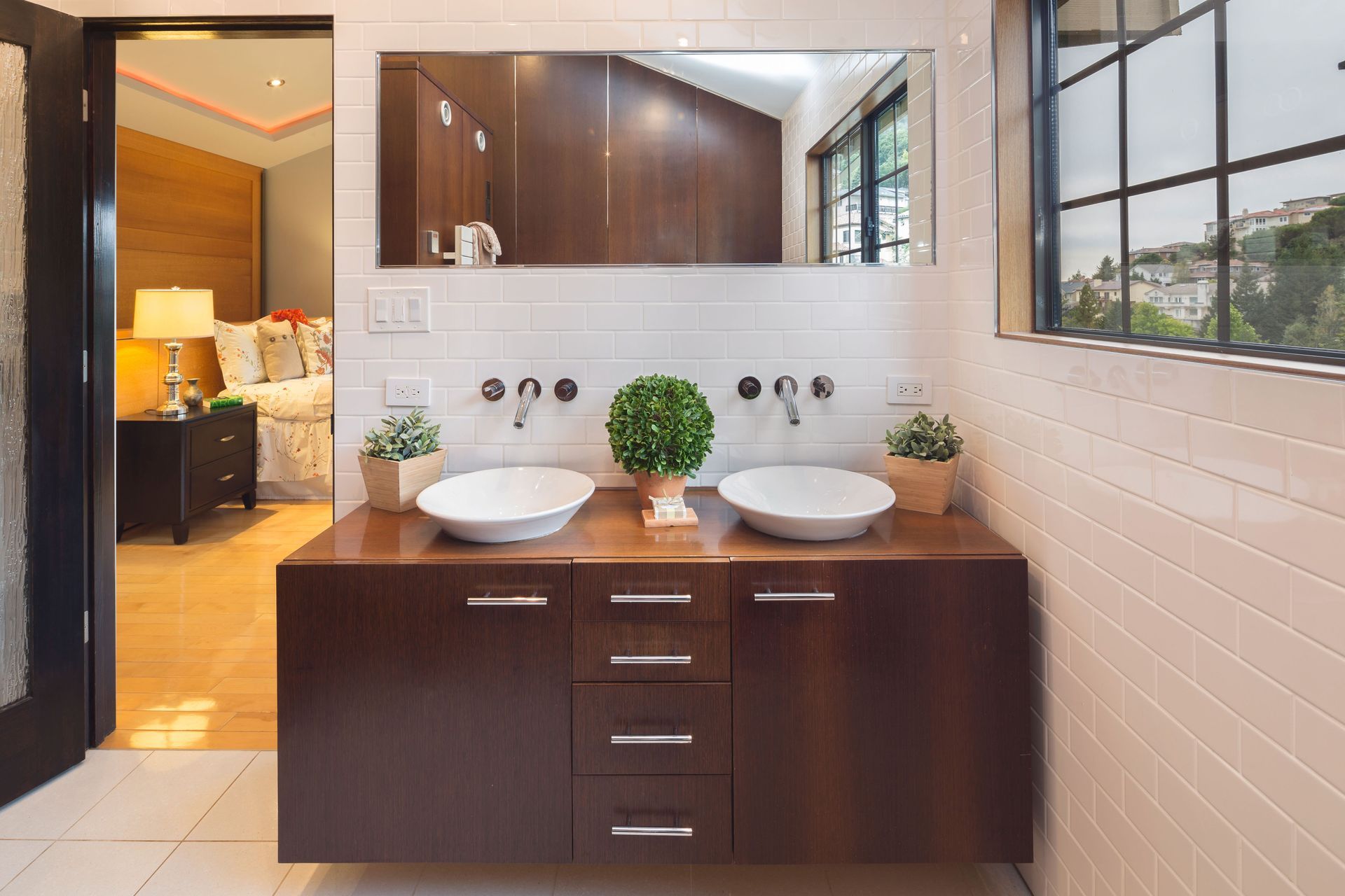 Bathroom with double sinks, brown vanity, large mirror, and view of bedroom.