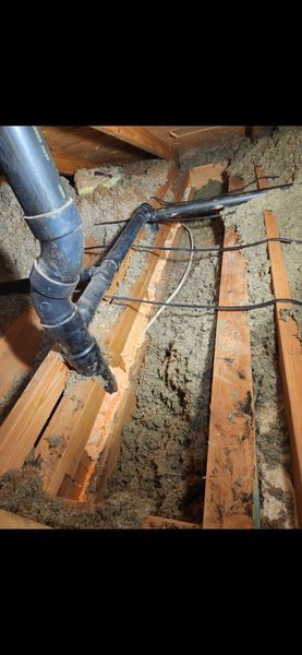 A black vacuum hose cleaning insulation in an attic. Wooden beams and loose insulation are visible.