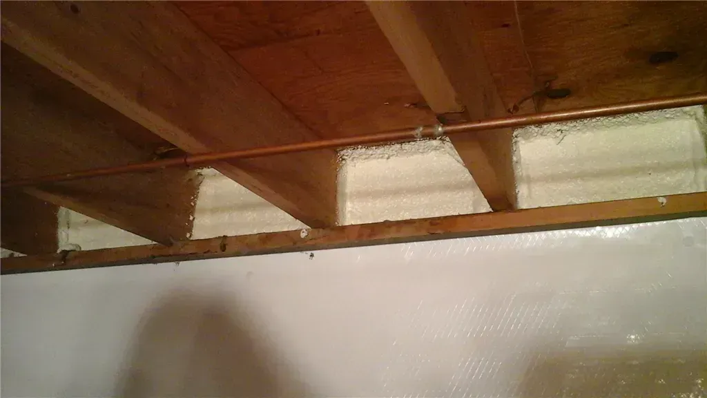 Foam insulation between floor joists, copper pipe, white painted wall.