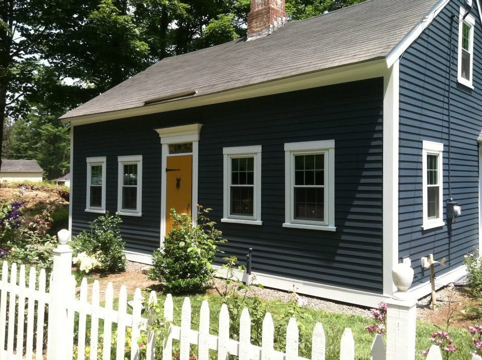 Dark blue cottage with white trim, orange front door, and white picket fence in a garden