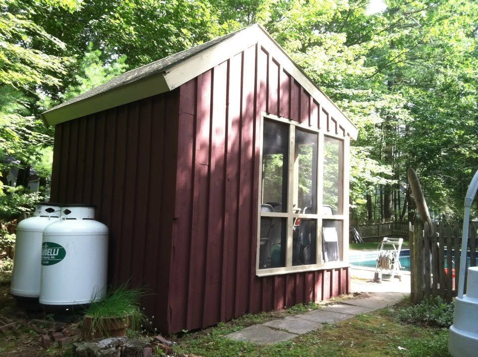 Small red garden shed with white trim beside a patio, surrounded by trees and a propane tank