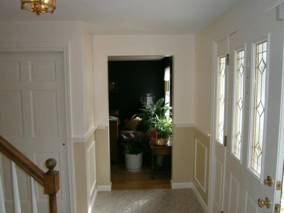 Hallway with white walls, wooden stairs, double doors, and a doorway to a dark room with plants.